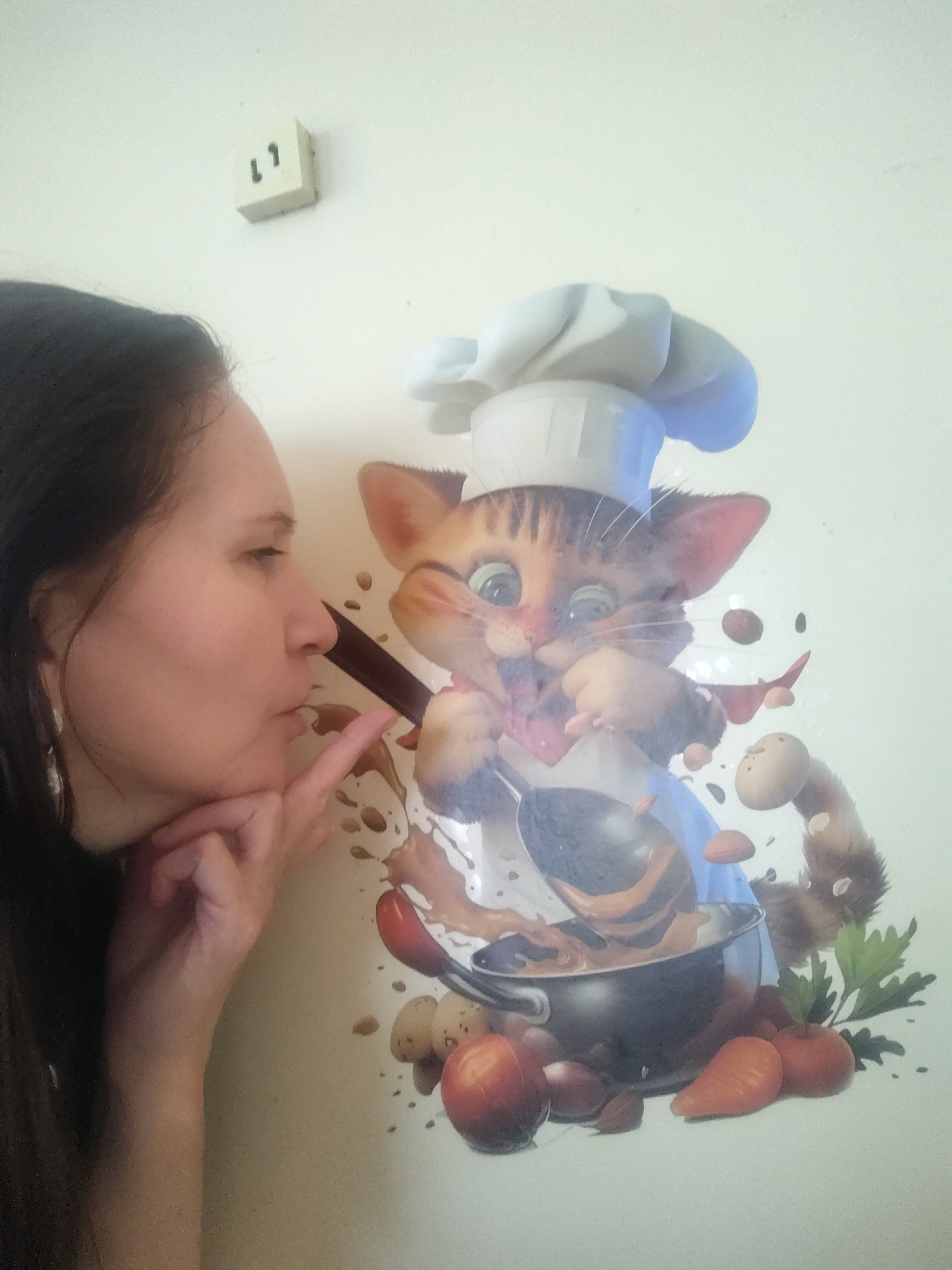 A woman stands to the left of a cartoon depiction of a cat chef. The woman has long brown hair, and is wearing earrings. She is holding her chin with her right hand, and extending her index finger toward the cartoon cat's spoon. The cat in the cartoon is wearing a chef's hat and a blue apron. It is stirring a pot with a spoon. Vegetables surround the pot. Above the cat cartoon, mounted on the wall is a white wall electrical outlet. The city is Sarapul, Russia.
