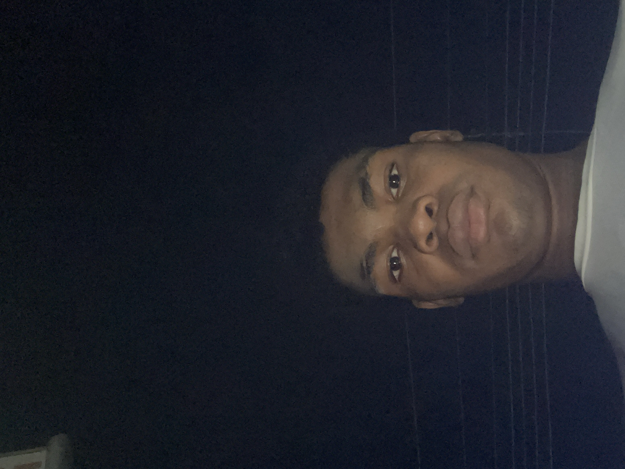 A close-up image of a dark-skinned person's face against a dark background, possibly taken at night. The individual has short, dark hair, visible eyes, nose, and lips. Part of a light-colored shirt is visible on the right side. The background appears dark and indistinct, possibly indicating an outdoor location at night in Ado, Nigeria, but there is no visible landmarks to confirm the city. No interactions or actions are apparent. The image's orientation is rotated sideways.