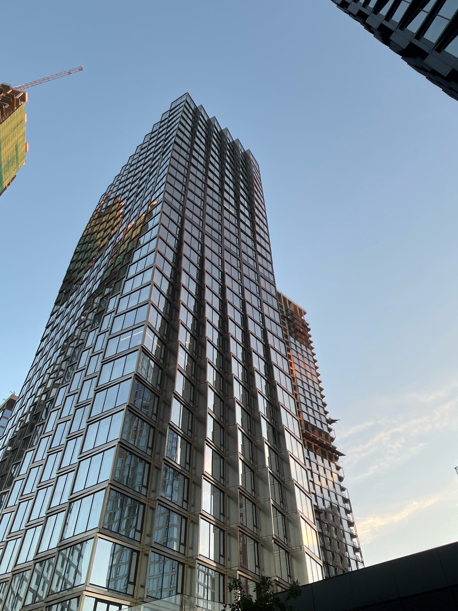 A low-angle shot shows a modern glass skyscraper under a clear blue sky in Moscow, Russia. The building's facade is composed of numerous rectangular glass panels, creating a reflective surface that mirrors the sky and surrounding environment. In the background, another partially constructed building with a crane is visible, suggesting ongoing development in the area. The sun is setting, casting a warm glow on the buildings.