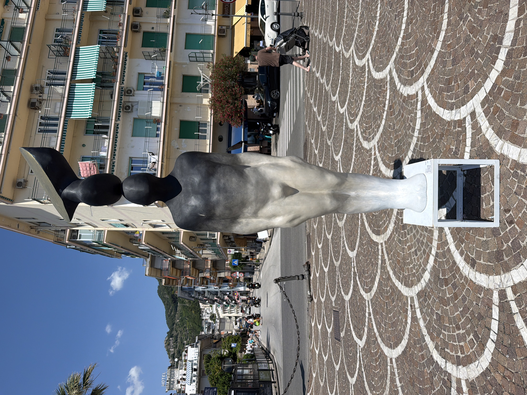 The image captures a sculpture in Maiori, Italy, set on a paved area with a distinct pattern of semi-circular white lines embedded in a brown stone base. The sculpture is predominantly in shades of grey and black, featuring a smooth body with rounded shapes, supported by a white rectangular pedestal. A person wearing a brown shirt is pushing a baby stroller on the sidewalk behind the sculpture. Buildings with green-striped awnings stand along the street on the left side of the image. In the distance, a road leads into the town, with visible vehicles and people. The background includes a mountain partially covered in greenery, under a clear blue sky.