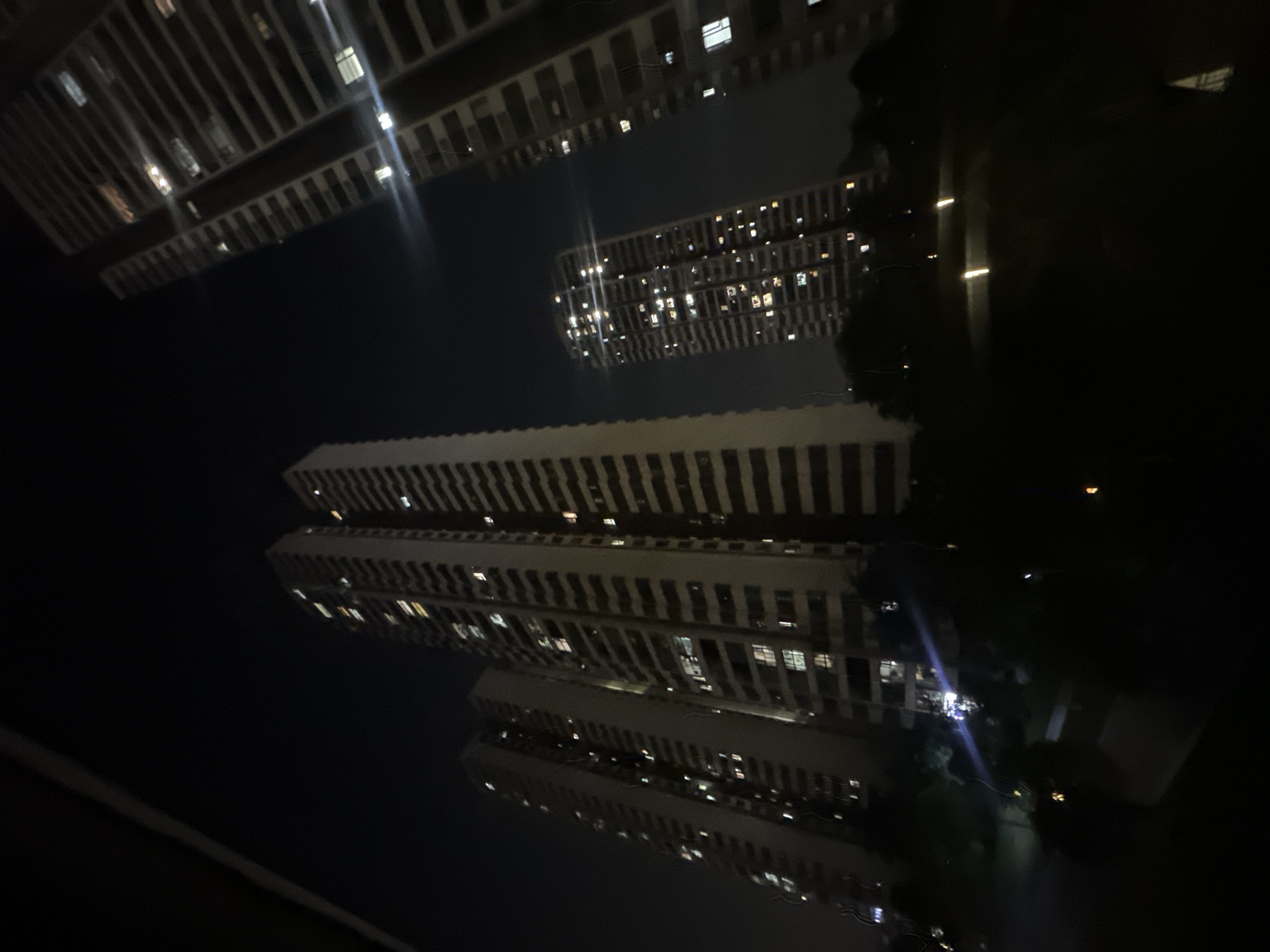 The image displays a night scene of multiple high-rise buildings in an urban environment. The photograph is oriented horizontally, showing tall, multi-story structures extending across the frame. These buildings feature numerous illuminated windows, creating patterns of light against the dark night sky. The most prominent structures appear as long, rectangular blocks.
A significant feature of the image is the clear and detailed reflections of these buildings, which occupy the top and bottom portions of the frame. These reflections are nearly perfect mirror images of the buildings' illuminated windows and architectural details, strongly suggesting the photograph was taken across a still body of water, possibly a canal, river, or large reflective surface like a wet street. Some faint, dark silhouettes, possibly trees or other structures, are vaguely visible between the buildings and their reflections in the darker areas.
The time of day is clearly nighttime, with artificial lights from the buildings being the primary light source. The overall atmosphere is dark and serene. There are no discernible people, specific activities, or visible text. The setting is the Chongqing High-tech Zone in China, captured in its night-time stillness.