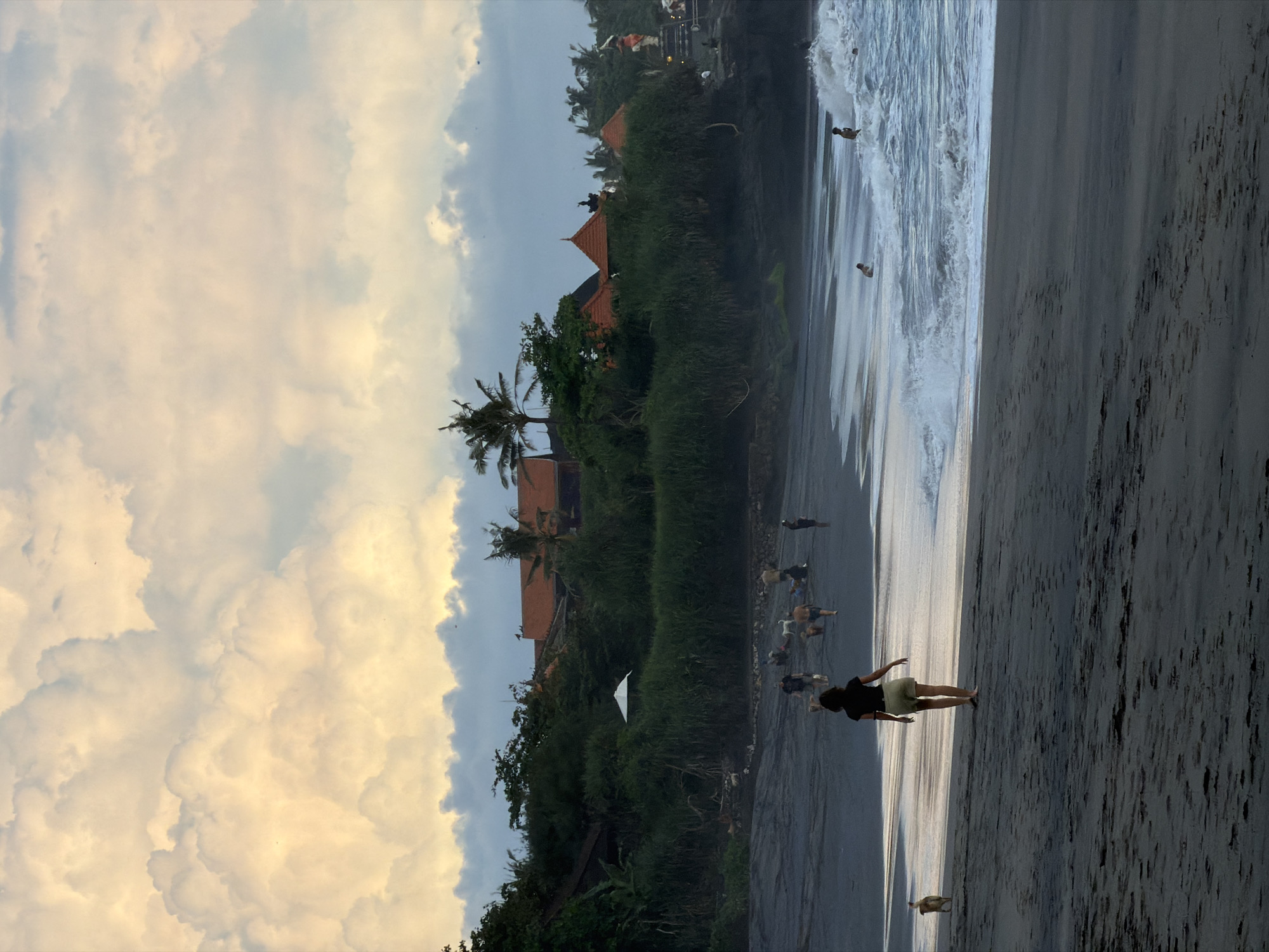 The image captures a coastal scene in Mengwi, Indonesia. On the left, lush tropical vegetation, including palm trees, covers the land, with glimpses of buildings featuring orange roofs. Above, the sky is filled with dense, puffy clouds. The beach is visible in the foreground. At the beach, there is a woman walking on the sand. Further down the shore are several people in the water. The waves are crashing, creating a dynamic interplay between the ocean and the shore. The overall atmosphere suggests a tropical location with a combination of natural and man-made elements.
