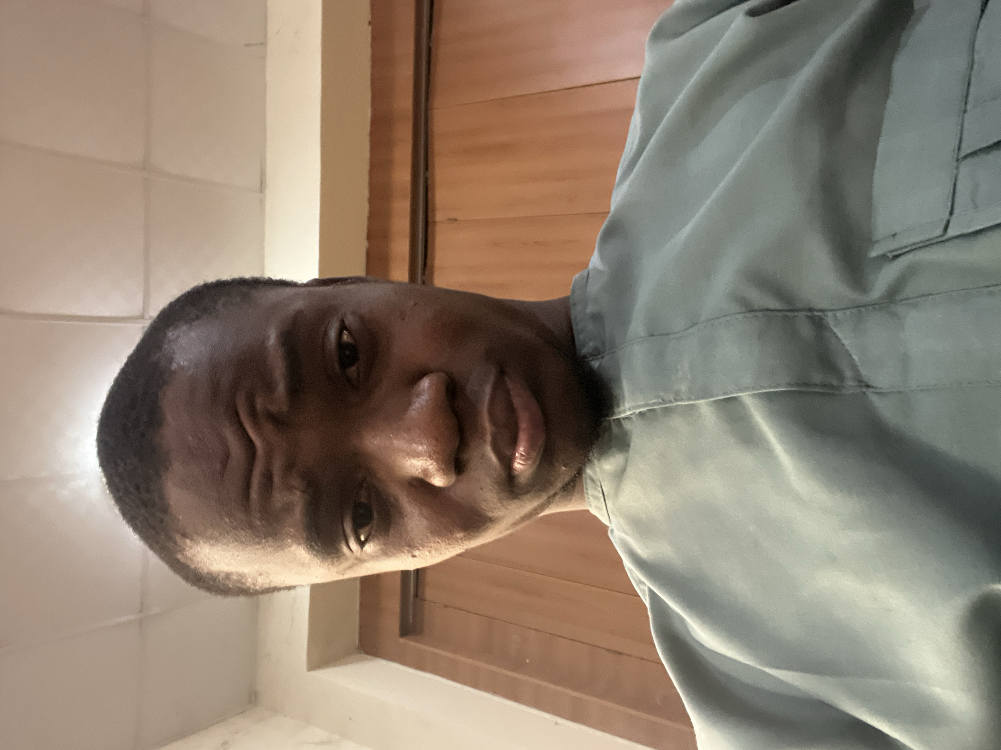 A medium close-up, slightly angled shot shows a dark-skinned man wearing a light-green or gray shirt with a visible pocket. He appears to be indoors, with a wooden door or panel behind him and a tiled or white wall visible on the left. The man's expression is somewhat serious, with furrowed brows. The image does not allow for confirmation of location details within Baganakwo, Nigeria.