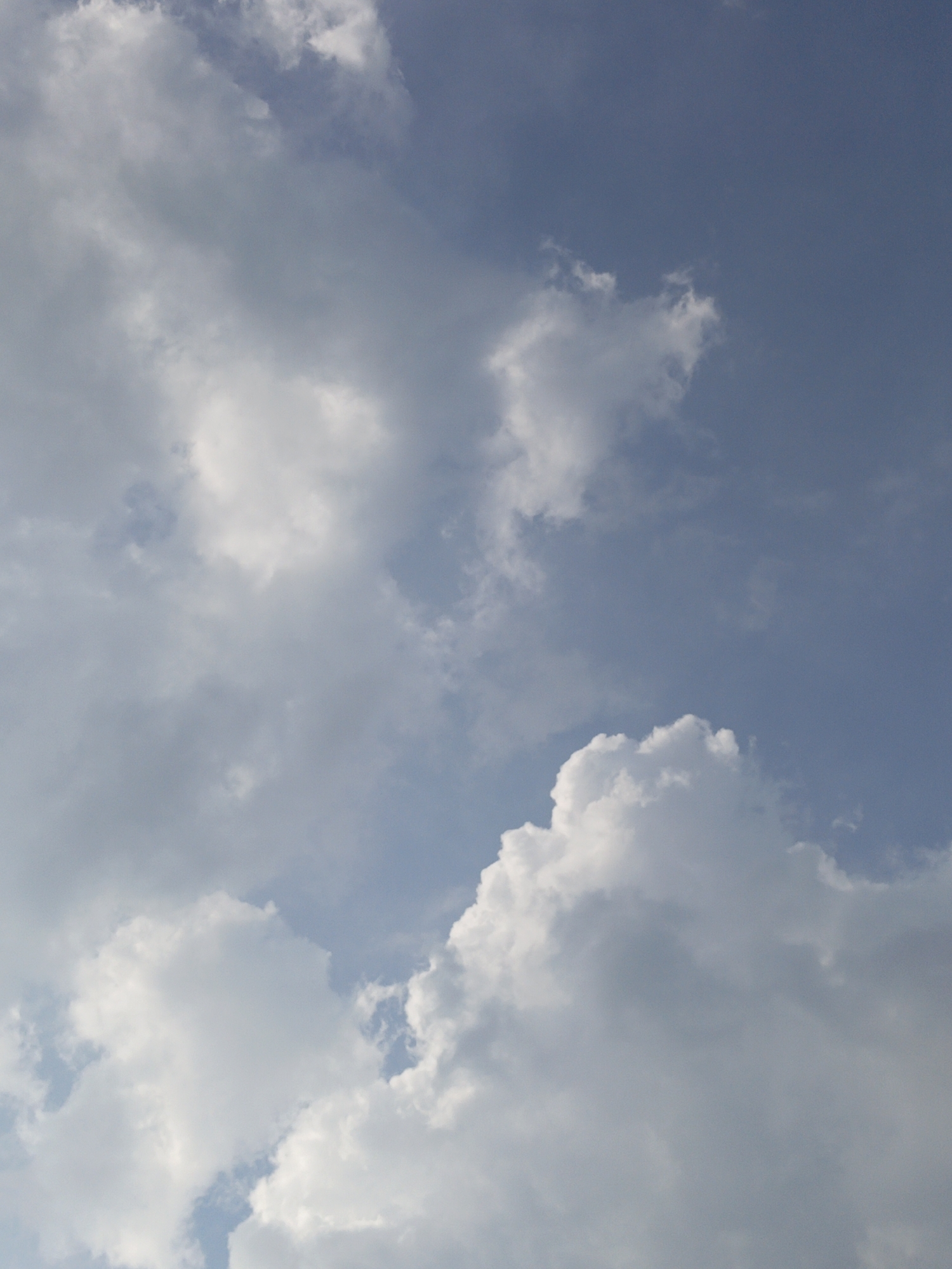 The image depicts a clear blue sky with scattered white and gray clouds. The clouds vary in density and shape, with some appearing puffy and illuminated by sunlight, while others are more diffuse and shadowed. The sky occupies the majority of the frame, suggesting an outdoor scene. There is no visible indication of the city of Gedung Nyapah, Indonesia, nor are there any people, subjects, or objects present in the image. The scene is a natural outdoor environment.
