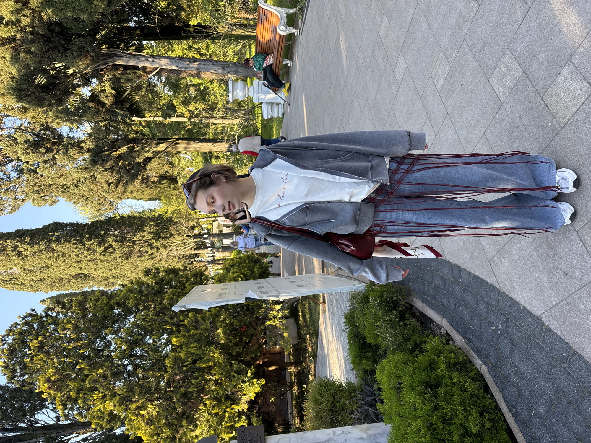 The image captures a young woman standing on a paved surface in a park-like setting in the city of Unknown, Unknown. She is wearing jeans with red strings, a white t-shirt, and a grey hoodie. She has sunglasses on her head and is carrying a red bag. A sign is seen in front of the woman. A bench is present in the background where another person sits. Trees surround the area, and sunlight filters through their leaves, creating a bright outdoor environment.
