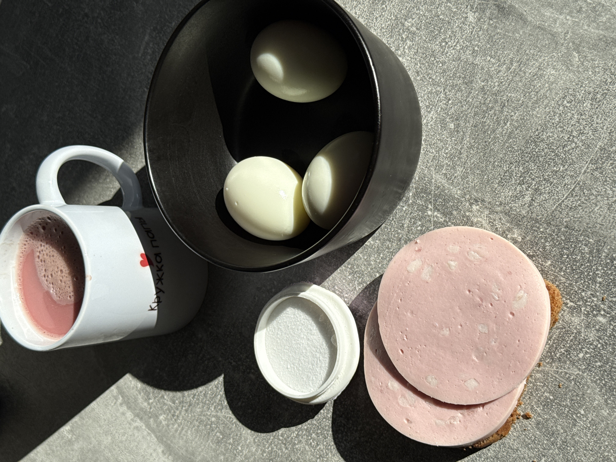 A breakfast scene is captured on a grey, textured surface under strong directional lighting, casting distinct shadows. In the upper left, a white ceramic mug, partially obscured by shadow, contains a pinkish beverage with froth. Text and a small red heart are visible on the mug. To the right of the mug is a dark, round bowl holding three peeled, hard-boiled eggs. In the lower left, two circular slices of pink sausage are placed on a piece of dark bread. A small, white lid with a textured surface sits next to the sausage. The scene appears to be an indoor setting, possibly a kitchen or dining area. There is no specific indication of the location being in Lyubertsy, Russia, beyond the user's prompt.