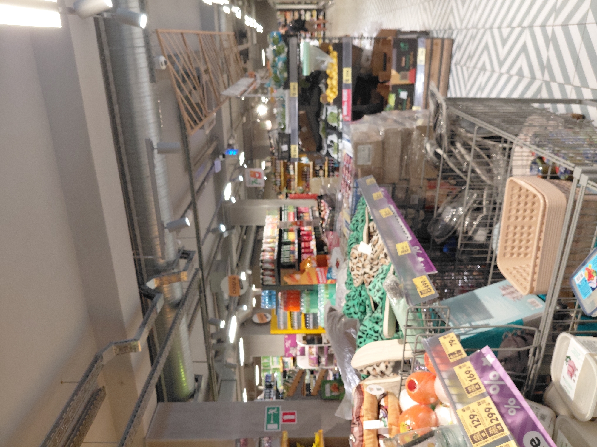 This is a medium-shot, indoor photograph taken at a diagonal angle within a supermarket in Novosibirsk, Russia. The lighting is bright and appears to be artificial, emanating from ceiling fixtures. The aisle is filled with various products, predominantly on shelves and in shopping carts.
In the foreground, on the left, is a shopping cart containing several items. These include a beige plastic basket, a clear plastic container with a light blue lid, and several other items obscured by the basket and the overall blurriness of the image. A portion of a price tag with the number "129" is visible on a purple strip. Further to the left, other products are visible, including what appear to be orange spherical objects and a clear bottle with a yellow label.
The middle ground of the image displays more shopping carts filled with merchandise. One cart contains a large amount of green fabric or material with a leopard-print pattern, and what appear to be slippers. Another shopping cart in the middle distance holds several boxes stacked and wrapped in plastic.
The background shows rows of shelves stocked with various goods, including bottled drinks, canned items, and packaged products. A sign with the word "СКИДКА" (meaning "discount" in Russian) is visible hanging from the ceiling. In the far right background, a person wearing a dark shirt and light-colored pants is walking away from the camera. The floor is tiled with a pattern of light grey and white diagonal lines. The overall impression is a busy supermarket environment.