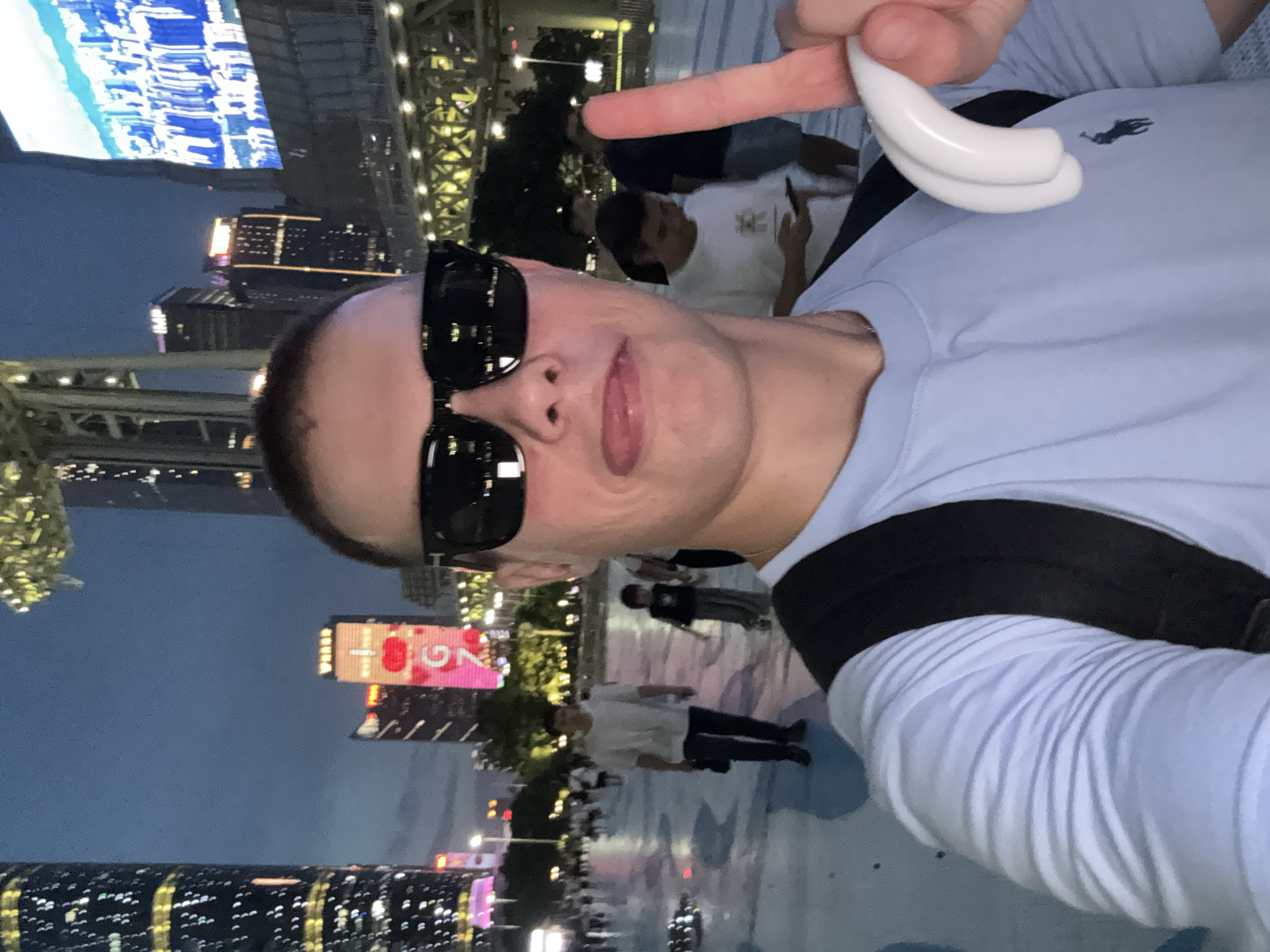 The image shows a person taking a selfie at night in a city. The person is wearing sunglasses and a light blue long-sleeved shirt with a dark blue logo. They are holding up a white object to the camera with their right hand. In the background, there are tall buildings with illuminated windows, some of which are lit with colorful lights. There is also a large screen displaying a blue and white pattern. Other people are visible in the background, appearing to be walking around. The sky is dark, indicating it is nighttime. The overall scene suggests an urban environment, likely a commercial or entertainment district. The presence of illuminated buildings and a large screen hints at a vibrant city at night. The image appears to be taken outdoors in what looks like a pedestrian area. There is text visible on a building in the background that appears to be in Chinese, with a prominent "26" visible. The person in the foreground has a neutral expression.