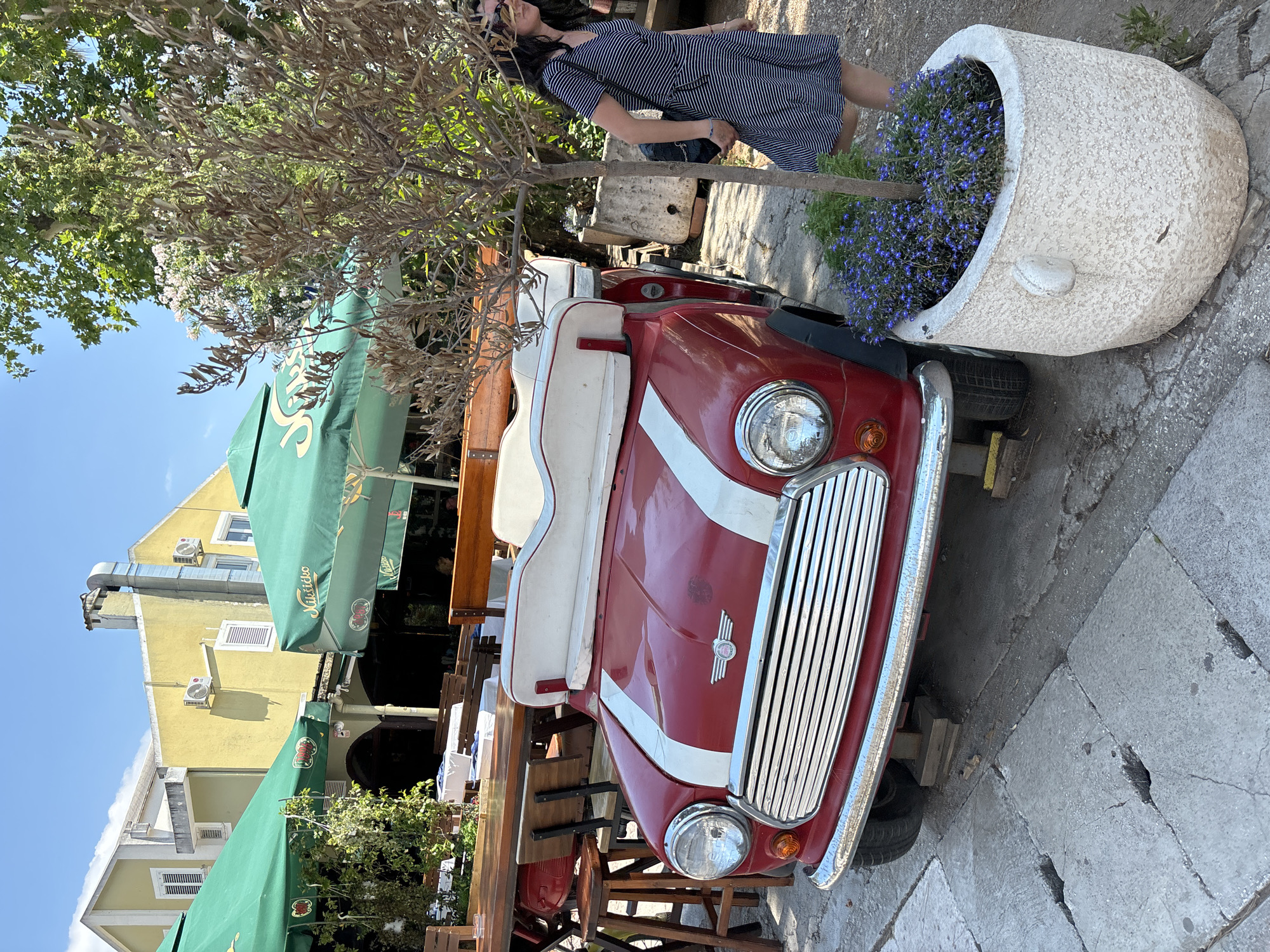 The image depicts an outdoor seating area of a restaurant in Budva, Montenegro. A red Mini Cooper convertible, possibly a decorative piece or a prop, is parked in front of a table and chairs. A woman in a striped dress walks past a planter filled with blue flowers and a small tree. Green umbrellas provide shade over the tables, and a yellow building with white shutters is visible in the background. The scene appears to be in a pedestrianized area, with cobblestone pavement.