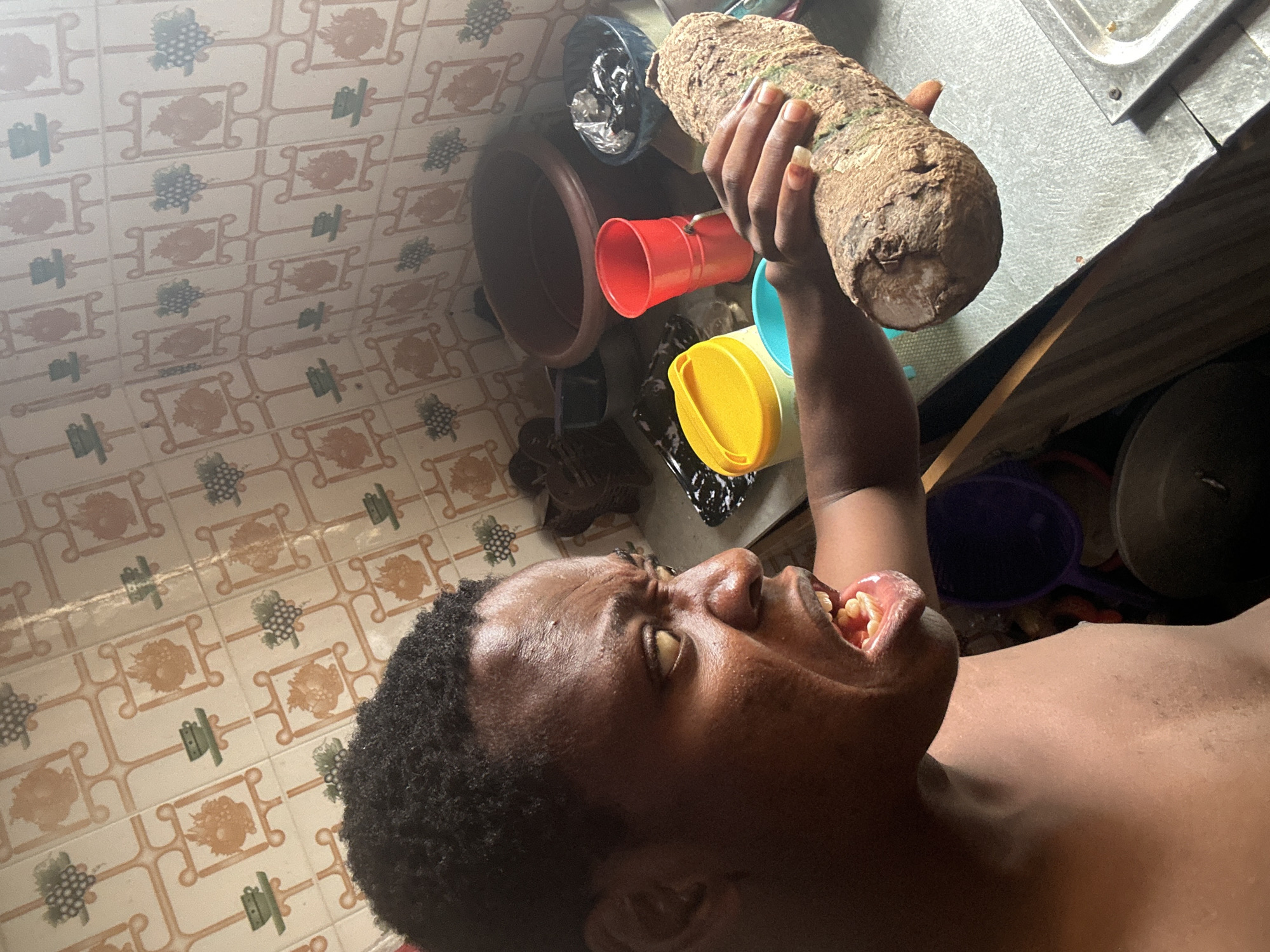 The image depicts a person with dark skin and curly black hair holding a large, brown, root vegetable up to their open mouth. The person's head is tilted back, and their eyes are visible. Several colorful plastic containers surround the root vegetable. The background includes a tiled floor and a stainless steel countertop. The location is Unknown, Unknown.