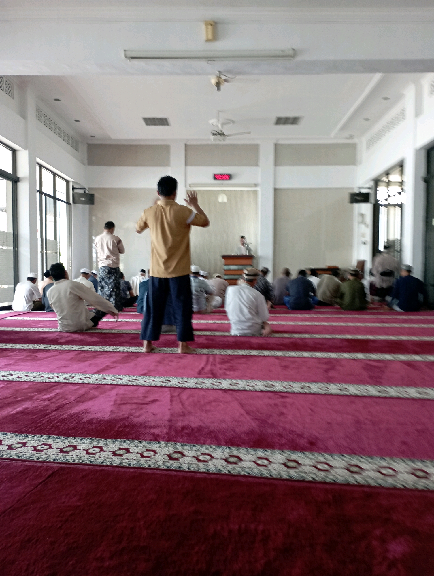 This is a photograph taken inside a mosque in Malang, Indonesia, likely during a prayer session. The foreground is dominated by a vibrant pink carpet with decorative borders. In the middle ground, several men are visible, some standing and others in various prayer positions. Two men in the foreground are particularly prominent. One, wearing a mustard-yellow shirt and dark pants, stands with his back to the camera, facing towards the front of the mosque. He appears to be in a standing position, possibly leading the prayer or performing a congregational prayer. Another man, wearing a light-colored shirt and patterned pants, is seated on the carpet.
In the background, more men are seated on the carpet, facing the same direction as the man in the mustard shirt. The mosque interior features white walls and a high ceiling with fluorescent lighting. Large windows are visible on the left side of the frame, offering a glimpse of the outside. A digital clock displaying red digits is mounted on a wall in the distance. The overall atmosphere suggests a religious gathering.