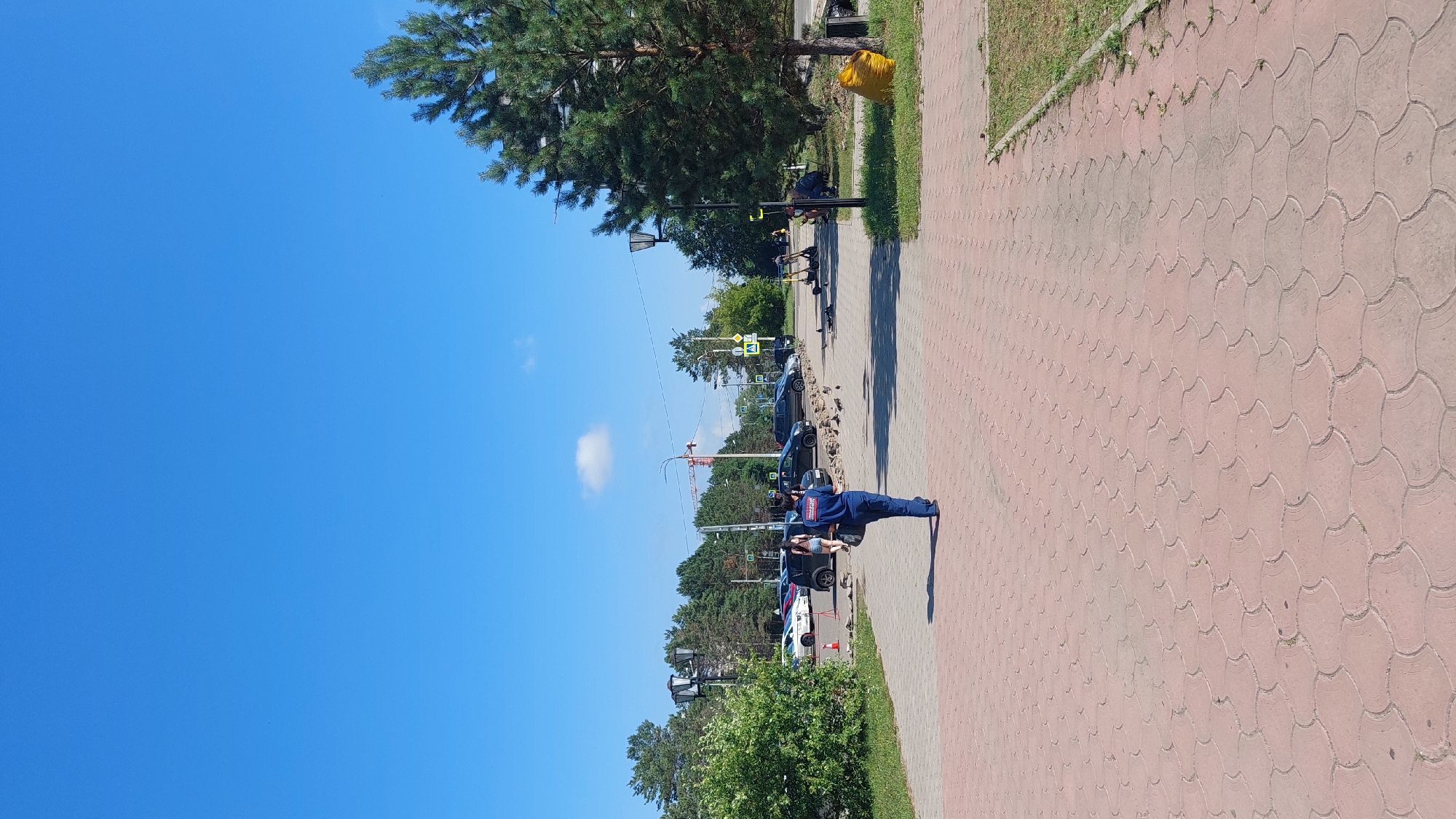 A bright, sunny day is depicted on a paved walkway in Komsomolsk-on-Amur, Russia. A person wearing a blue uniform walks away from the camera down the walkway, carrying a dark bag. In the background, there are several parked cars and a construction crane. Some pigeons are gathered on the ground near the cars. A large pine tree is visible on the right side of the image, and a yellow trash bag is at its base. The sky is clear blue with a few wispy clouds.