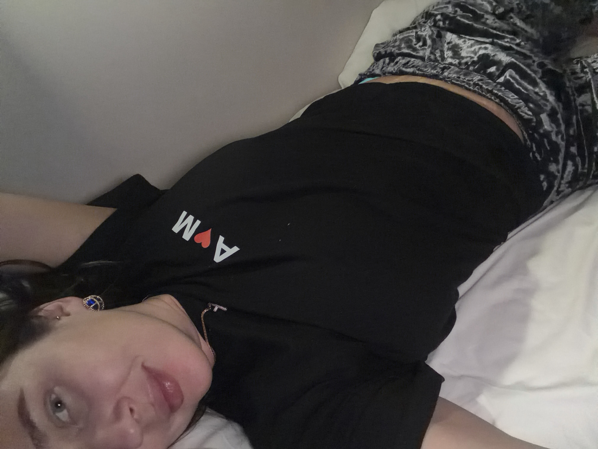 A woman lies on a white bed, wearing a black t-shirt with white letters and a red heart, and patterned pajama pants. The location cannot be determined.