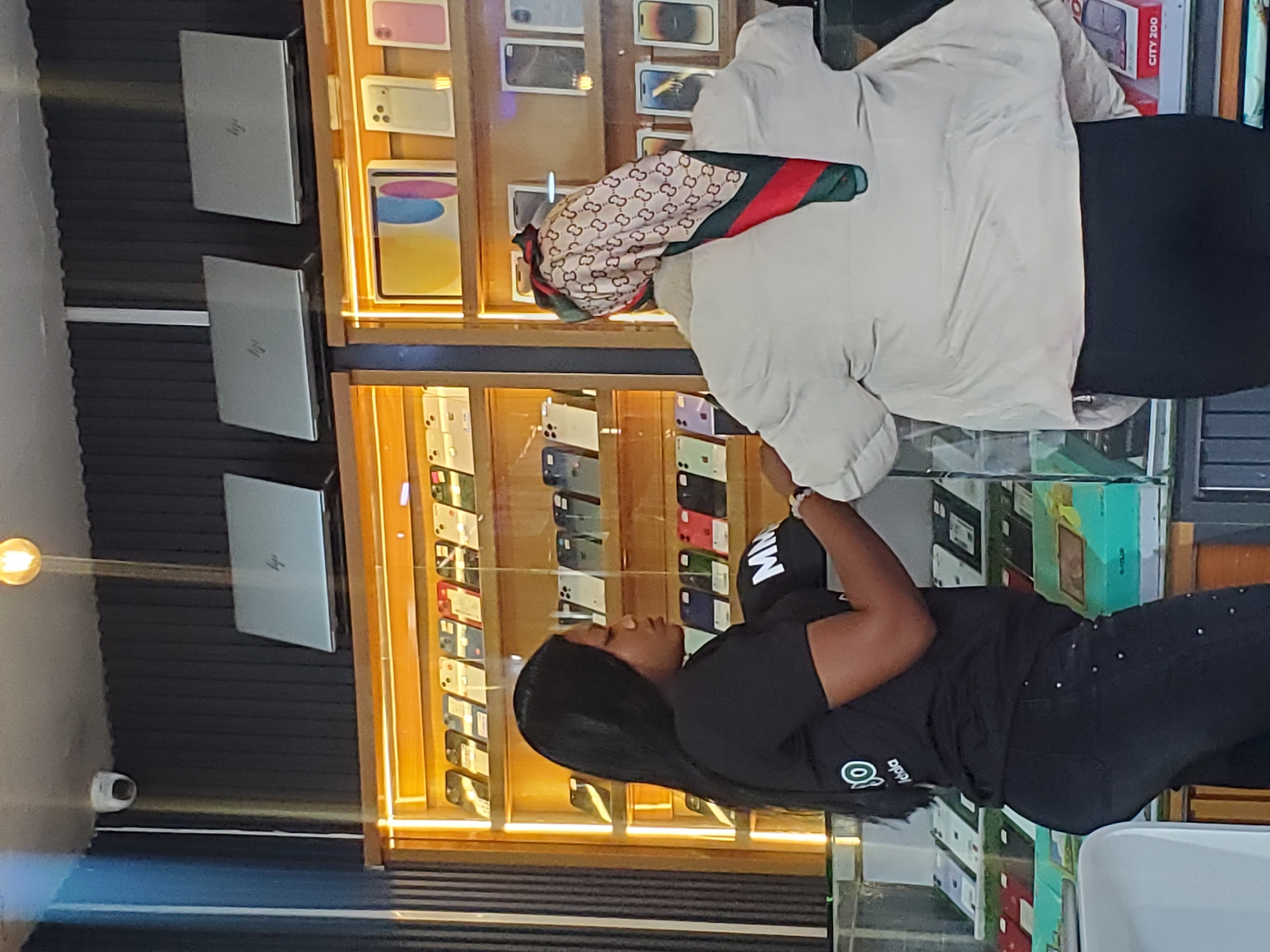 Two women are in what appears to be an electronics store in Jos, Nigeria. The woman in the foreground is wearing a black t-shirt with a logo and black pants, and her hair is long and dark. She is looking to her right. The woman behind her is wearing a light-colored jacket over a black garment and a scarf with a patterned design. She is also looking to her right, away from the camera. The store has shelves filled with electronic devices, possibly phones and laptops. There are also some boxes on display. A security camera is visible on a dark wall to the left. The lighting suggests it could be daytime, and there are no clear indications of the weather. The overall atmosphere appears casual, and the women do not display any strong emotions. The logo on the foreground woman's shirt is partially visible and appears to be green and white. There is also text on a red box in the background which reads "CITY 200".