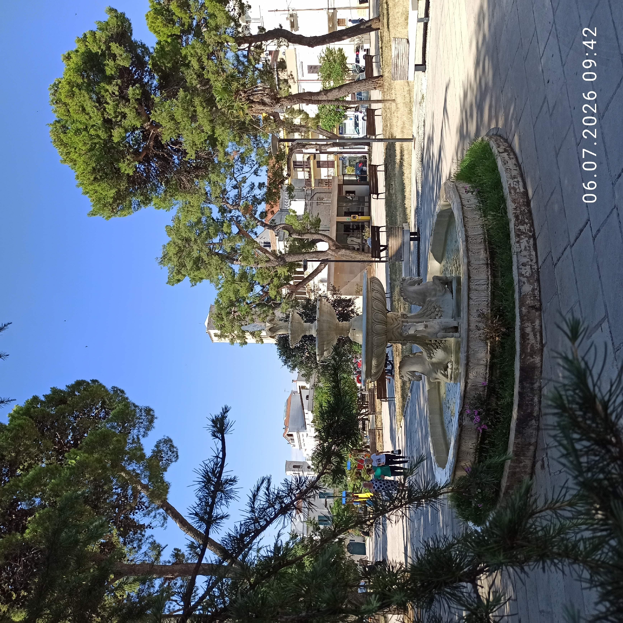 In Ruvo di Puglia, Italy, a sunlit public square features a three-tiered fountain with equine sculptures at its base. The fountain is surrounded by grass and situated on a paved area. Benches are scattered throughout the square, some occupied by people. Tall pine trees with lush green foliage frame the scene, casting shadows on the ground. In the background, buildings, including a tower with arched windows, and a playground are visible under a clear blue sky. The timestamp "06.07.2026 09:42" is present in the lower right corner.