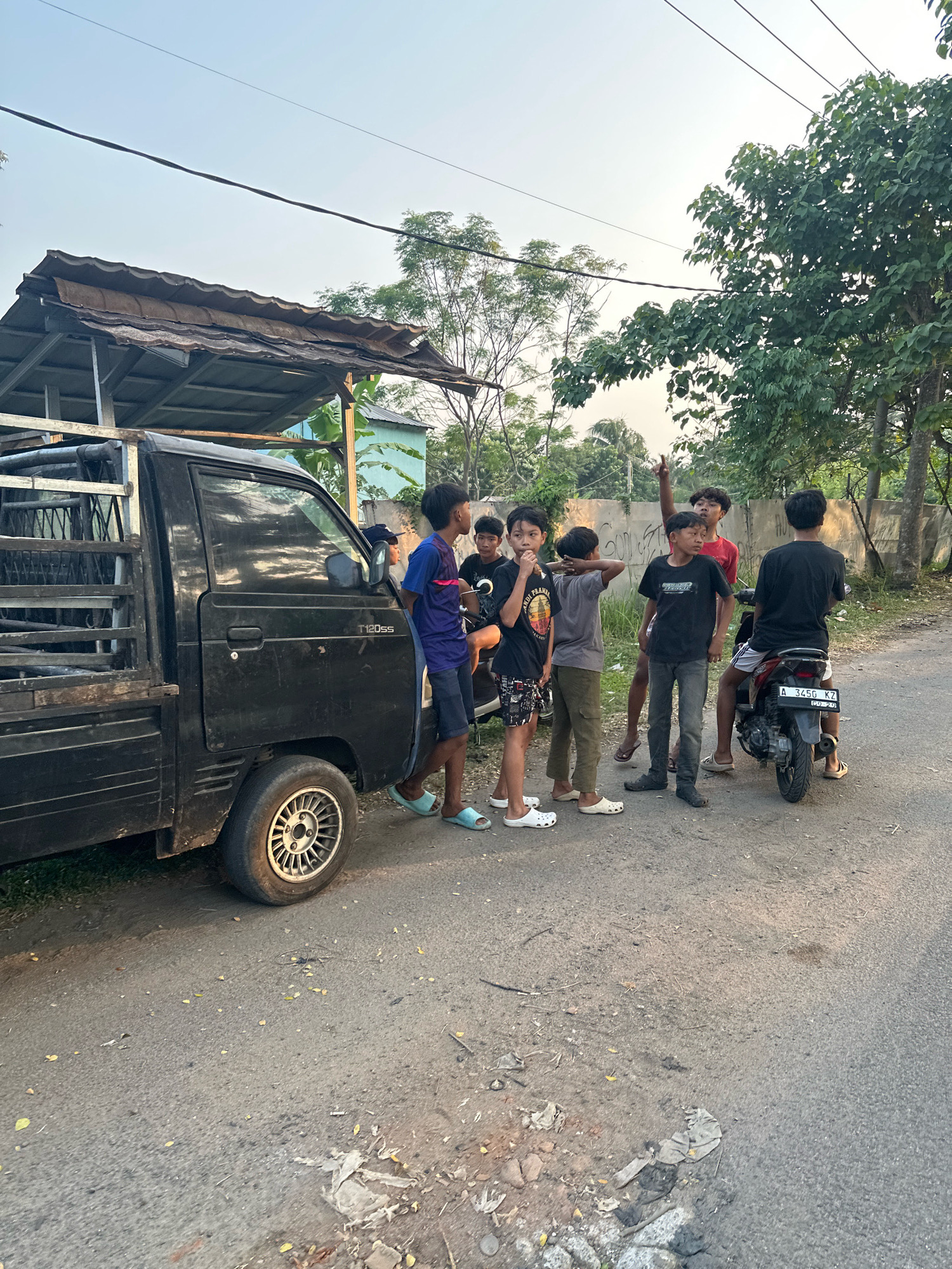 In Saruni, Indonesia, a group of young males gathers on a rural road beside a black pickup truck and a motorcycle. The truck, parked on the left, has a weathered, corrugated metal roof extending over its bed. One young man, wearing a blue shirt and dark shorts, leans against the truck. Another individual, in a red shirt, stands in the background, gesturing with his right hand raised. Several other boys, dressed in casual attire, are positioned around the truck and near a motorcycle on the right side of the road. The motorcycle, bearing the license plate A 3456 KZ, is occupied by one of the youths. The environment is characterized by a paved road with scattered leaves and debris, bordered by trees and vegetation. A concrete wall is visible in the background, along with a light blue building peeking through the trees. The sky above is clear and pale, suggesting it is either early morning or late afternoon.