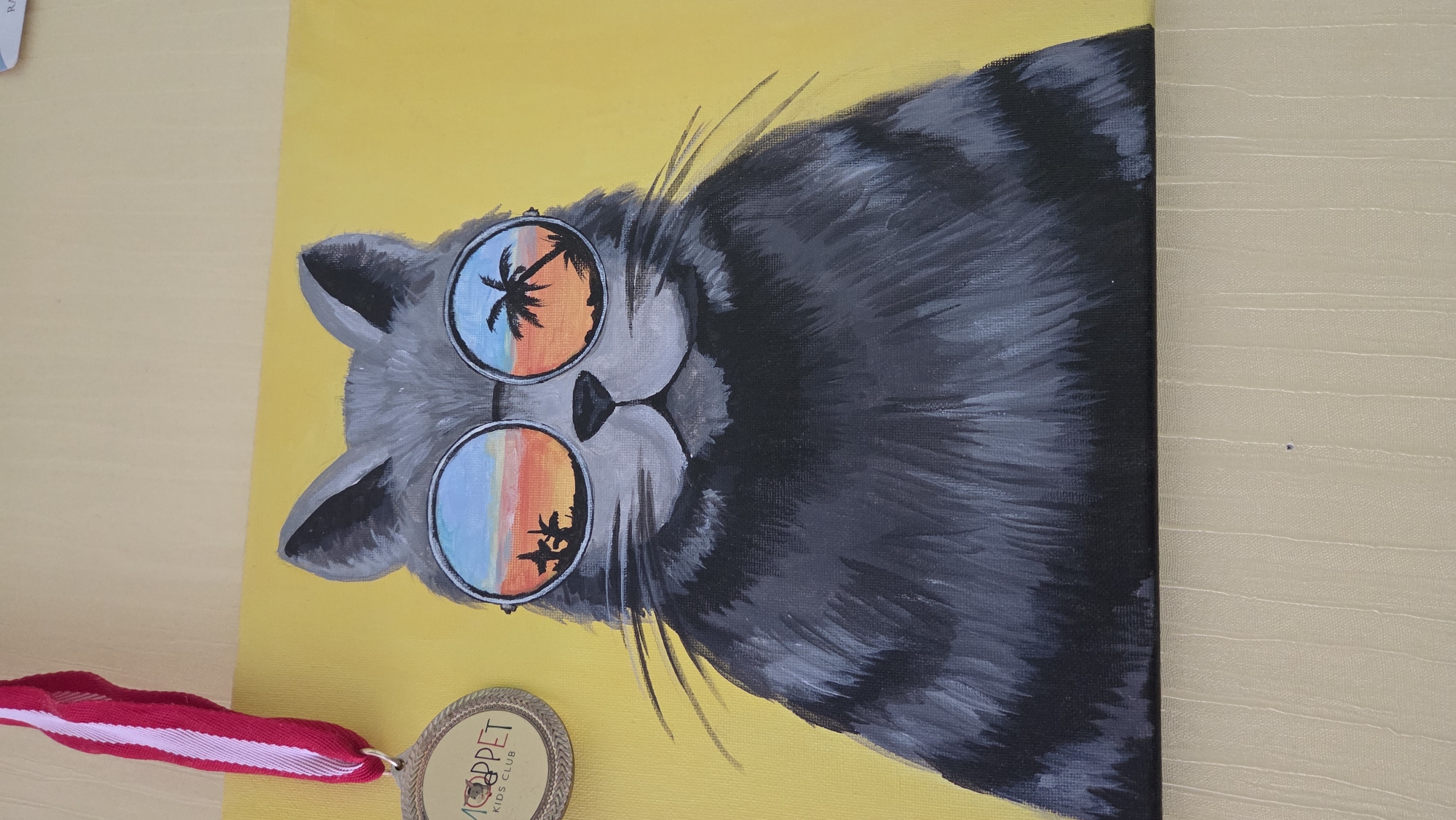 The image depicts a painting of a gray tabby cat wearing round sunglasses. The reflection in the sunglasses shows a tropical beach scene. The background of the painting is yellow. A medal with a red and white ribbon is placed next to the painting. The medal features the text "MOPPET KIDS CLUB". I have no information about the location of the painting being in Chaikovsky, Russia.
