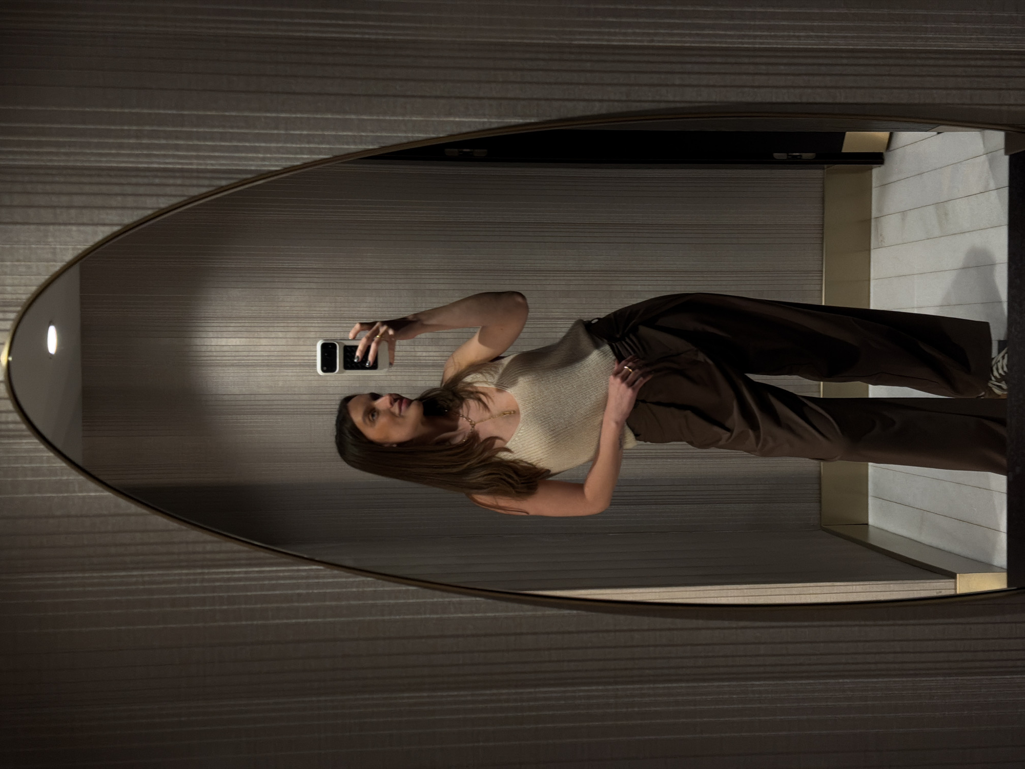 A woman is taking a mirror selfie in an oval mirror. She is wearing a light-colored ribbed tank top and brown wide-leg pants. The location appears to be an indoor space with horizontally textured walls and a vertical wall with light-colored panels and gold trim visible in the reflection. It is not possible to confirm the location in Budva, Montenegro.