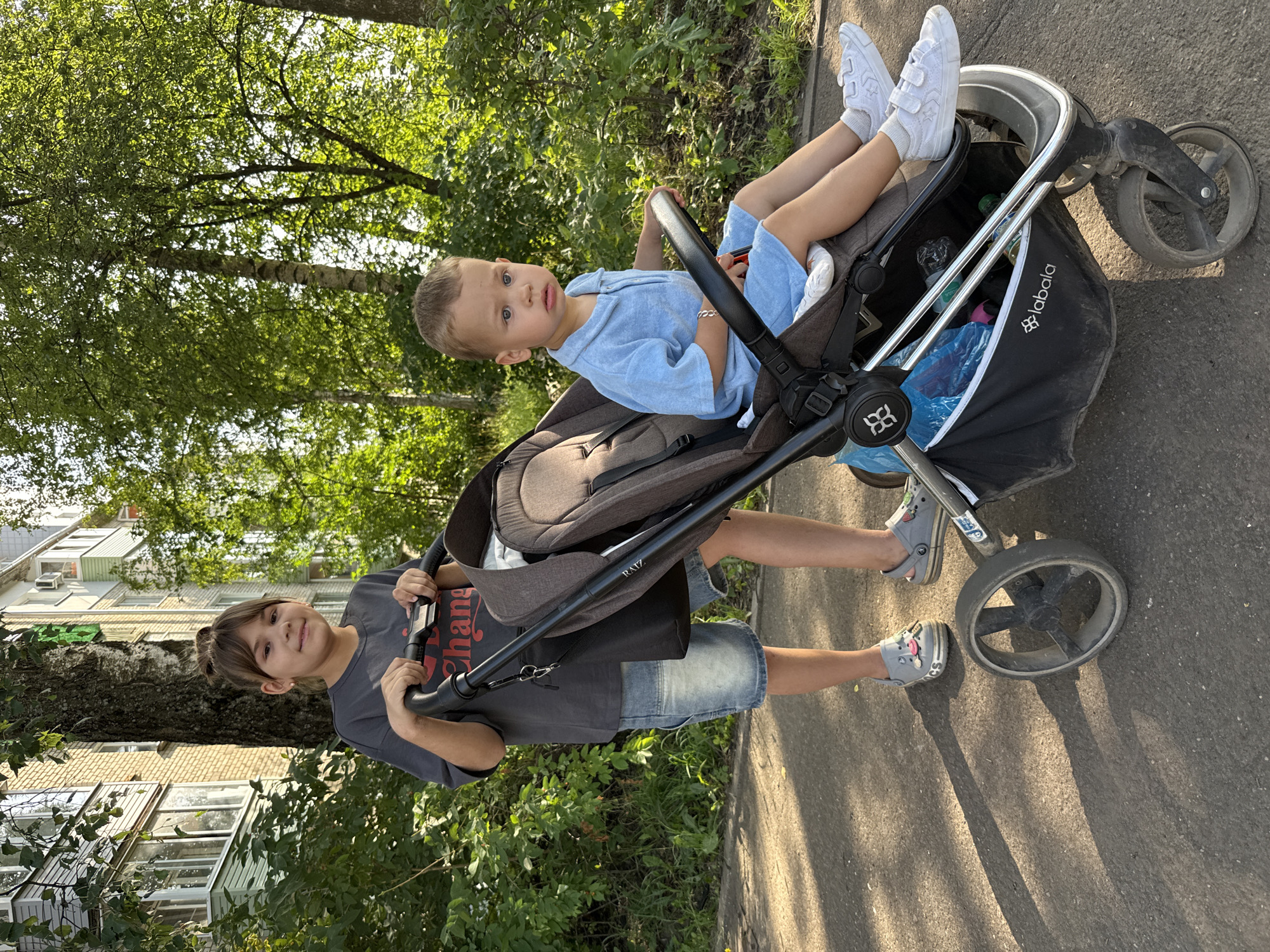 A young girl with brown hair in a bun, wearing a gray t-shirt and denim shorts, pushes a stroller. A young boy in a blue outfit sits in the stroller. They are on a paved path outdoors, with trees and a building in the background. The girl is looking at the camera with a smile. The boy is looking away from the camera with a neutral expression. The scene appears to be in a residential area.
