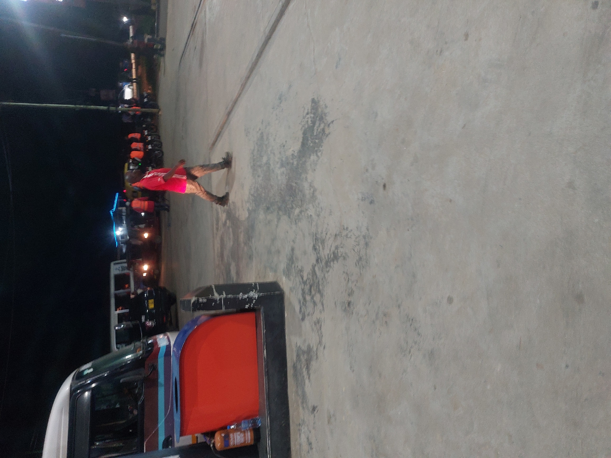 A poorly lit outdoor scene depicts a man wearing a red shirt, beige pants, and bare feet walking on a gray concrete surface. Behind him are parked trucks and buses, and people standing around the vehicles. An overhead wire is supported by a utility pole and streetlights are visible in the background. The scene appears to be in Dar es-Salaam, Tanzania, with a concrete gas or fuel pump structure present.