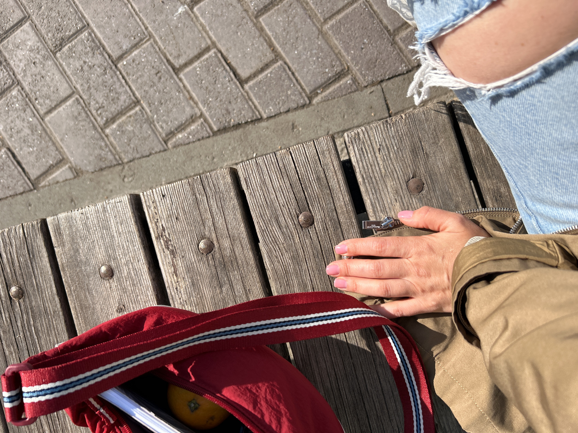 The image captures an overhead, close-up perspective of a person sitting outdoors on a sunny day in Chaikovsky, Russia. The scene is dominated by natural textures: weathered wooden planks forming a bench or boardwalk, and grey rectangular stone pavers of a sidewalk.
The subject's right leg, clad in light blue distressed jeans with a prominent rip on the thigh, is visible on the right side of the frame. Their right hand, with light pink painted nails, rests on a beige or khaki-colored jacket, near a metallic zipper pull which has some indistinct, unreadable text.
Next to the person is a bright red fabric bag. The bag features a distinctive strap with red, white, and blue stripes. Inside the open bag, an orange-colored fruit, possibly a tangerine or small orange, and a white-paged notebook or book are visible.
The strong, direct sunlight suggests it is daytime, casting clear shadows and highlighting the grain of the wooden planks and the texture of the pavers. The overall impression is one of a casual outdoor setting, possibly a park, promenade, or urban pedestrian area, with the person taking a moment of rest or waiting.