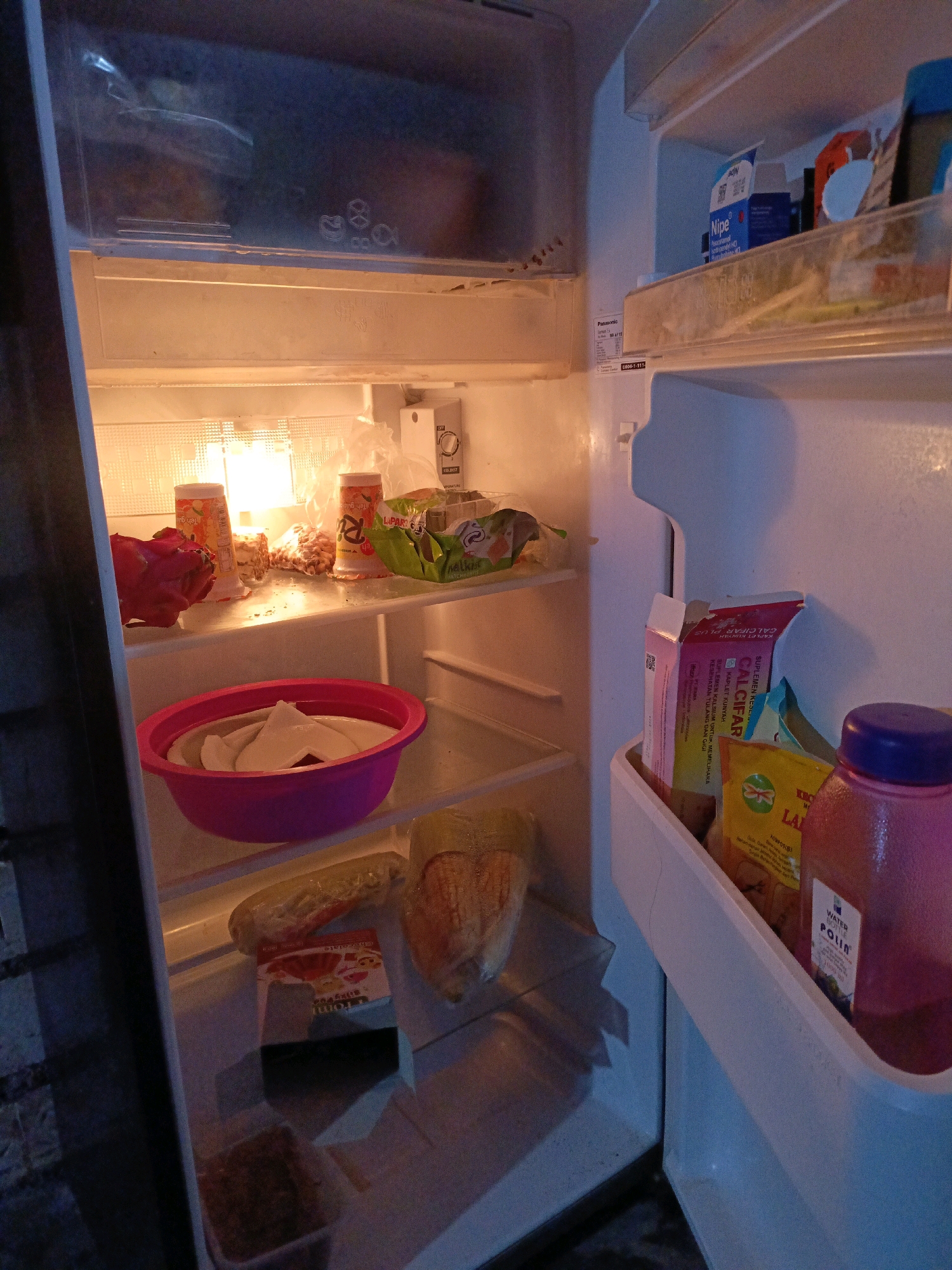 This is an interior, eye-level shot of a refrigerator, open on the right side. The refrigerator is lit from within by a bright light.
On the top shelf, to the left, there is a red dragon fruit. To the right of the dragon fruit are two orange cylindrical containers and some other items in plastic packaging. A clear plastic bag with items inside is on the far right.
The middle shelf has a bright pink bowl containing white paper. Below that, on the lower shelf, is a cucumber and a loaf of bread. A small box with a red and white label is on the shelf below the bread, and to its right is another item in a plastic bag.
The right side of the refrigerator features a door-mounted storage compartment. On the top shelf of this compartment are two boxes, one pink and one yellow, and a blue and white box. Further down, there is a large pink plastic bottle with a purple lid, labeled "WATER POLK".
The overall impression is that of a moderately stocked refrigerator. The location in the city of Jenu, Indonesia, is not discernible from the image.