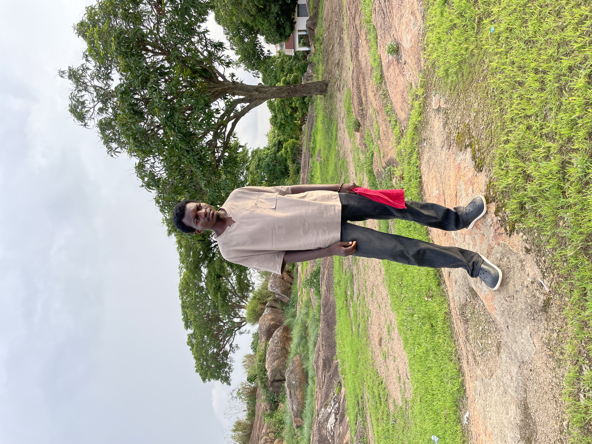 A man stands on a rock outcrop in Jos, Nigeria. He is wearing a tan shirt, black pants, and black shoes. He holds a red cloth. Behind him is a tree and a patch of grass and rocky landscape. The sky is overcast. A building can be seen in the background.