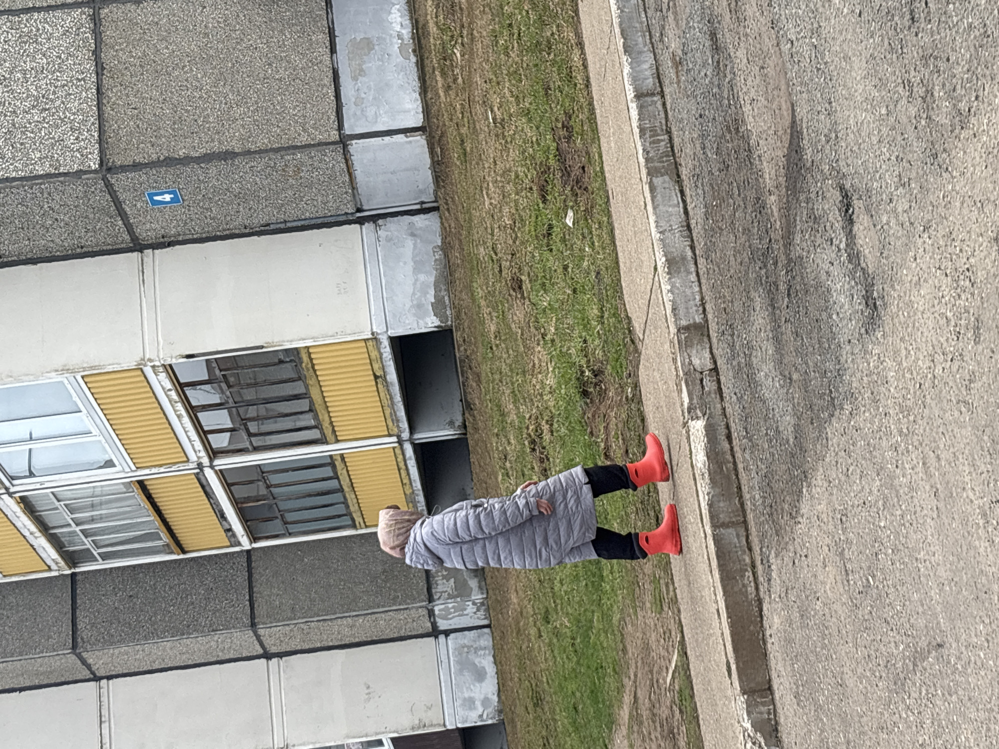 The image shows a woman walking on a sidewalk in Марковский, Russia. She is wearing a grey padded coat, black pants, and bright orange rubber boots. To her left is a grassy area, and beyond that is a grey concrete building with yellowish windows. A blue sign with a white arrow is visible on the building's facade. To her right is a grey concrete road.