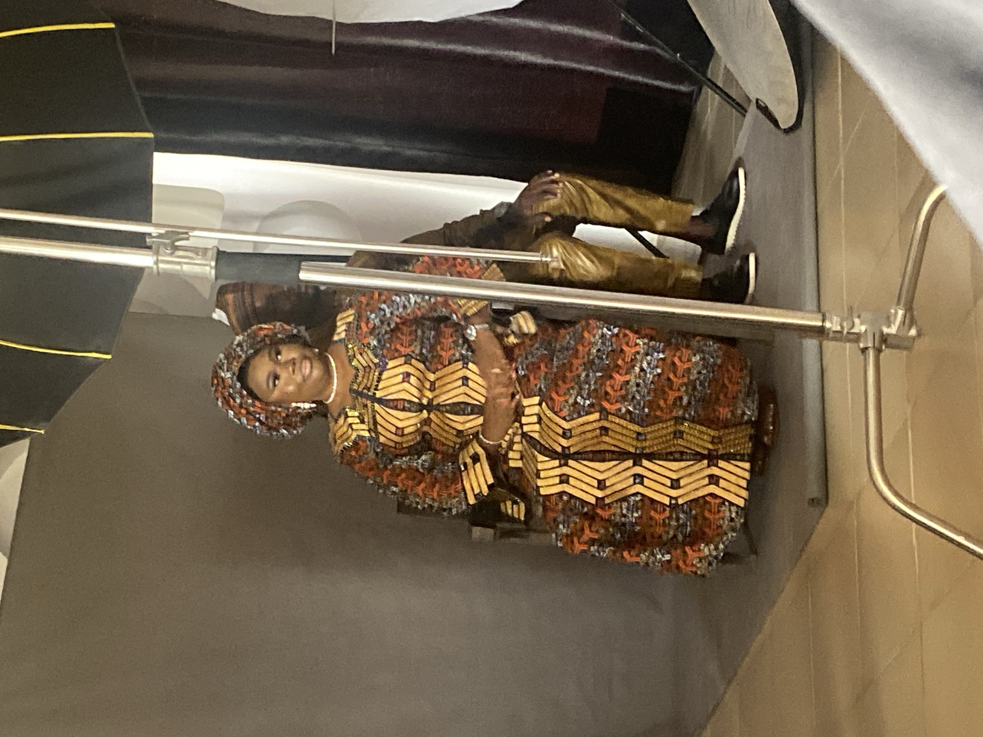 The image portrays an indoor scene, likely a photography studio or a prepared setup for a photoshoot, given the presence of lighting equipment. The central figure is a woman, radiating joy with a bright smile directed towards the viewer. She is elegantly dressed in a vibrant traditional African outfit, a long gown featuring intricate patterns in hues of orange, brown, and gold. The textile design incorporates geometric shapes, such as chevrons and stepped motifs, characteristic of West African aesthetics. Her ensemble is completed with a matching headwrap (gele), a white beaded necklace, earrings, and bracelets on her wrists, enhancing her festive appearance.
Partially visible in the background, behind the woman and the photography gear, is another individual, possibly a man, who appears to be seated. His lower body is visible, dressed in golden or light brown trousers and dark shoes.
The setting includes professional photography equipment: a black softbox with a yellow border is on the left, and several silver light stands or poles crisscross the frame, indicating an active photoshoot environment. The background comprises simple gray and beige walls, providing a clean backdrop for the portrait. The overall atmosphere conveys a sense of celebration and confidence, fitting the cultural context of Jos, Nigeria. No discernible text is visible in the image.