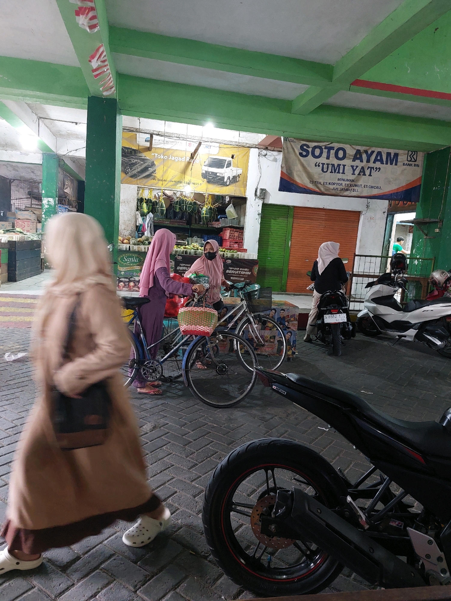 The image depicts a street scene in Sumuragung, Indonesia. In the foreground, a person wearing a tan abaya and white crocs is walking away from the camera, with their face obscured by motion blur. To the left, a bicycle with a woven basket is parked. In the middle ground, two women are interacting near a market stall. One woman, wearing a pink hijab and a dark face mask, is standing and talking to another woman who is seated on a motorcycle. This seated woman wears a pale pink hijab and a black top. Behind them, a large banner advertises "SOTO AYAM 'UMI YAT'". Further in the background, several motorcycles are parked, including a white scooter and a black motorcycle. The environment appears to be an open-air market with stalls and a covered ceiling supported by green pillars. The ground is paved with interlocking bricks.