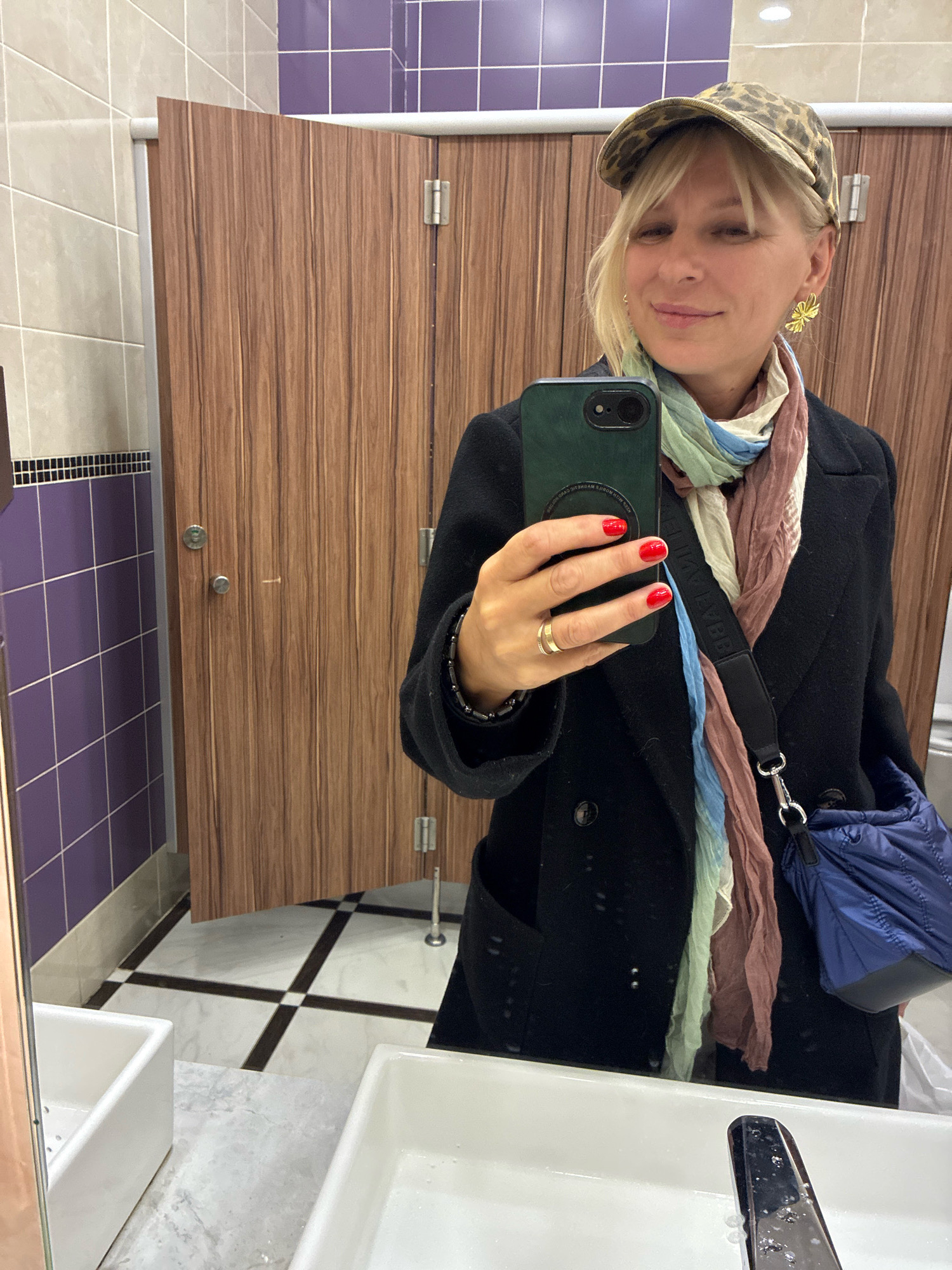 A woman is taking a selfie in a public restroom. She is wearing a black coat, a leopard print cap, and a multi-colored scarf. She has red nail polish and a gold ring on her left hand, which is holding a dark green smartphone. She is also wearing a gold butterfly earring on her right ear. A blue quilted bag is slung over her shoulder. The restroom has wooden stall doors, purple and beige tiled walls, and a black and white checkered floor. There are two white sinks in the foreground.