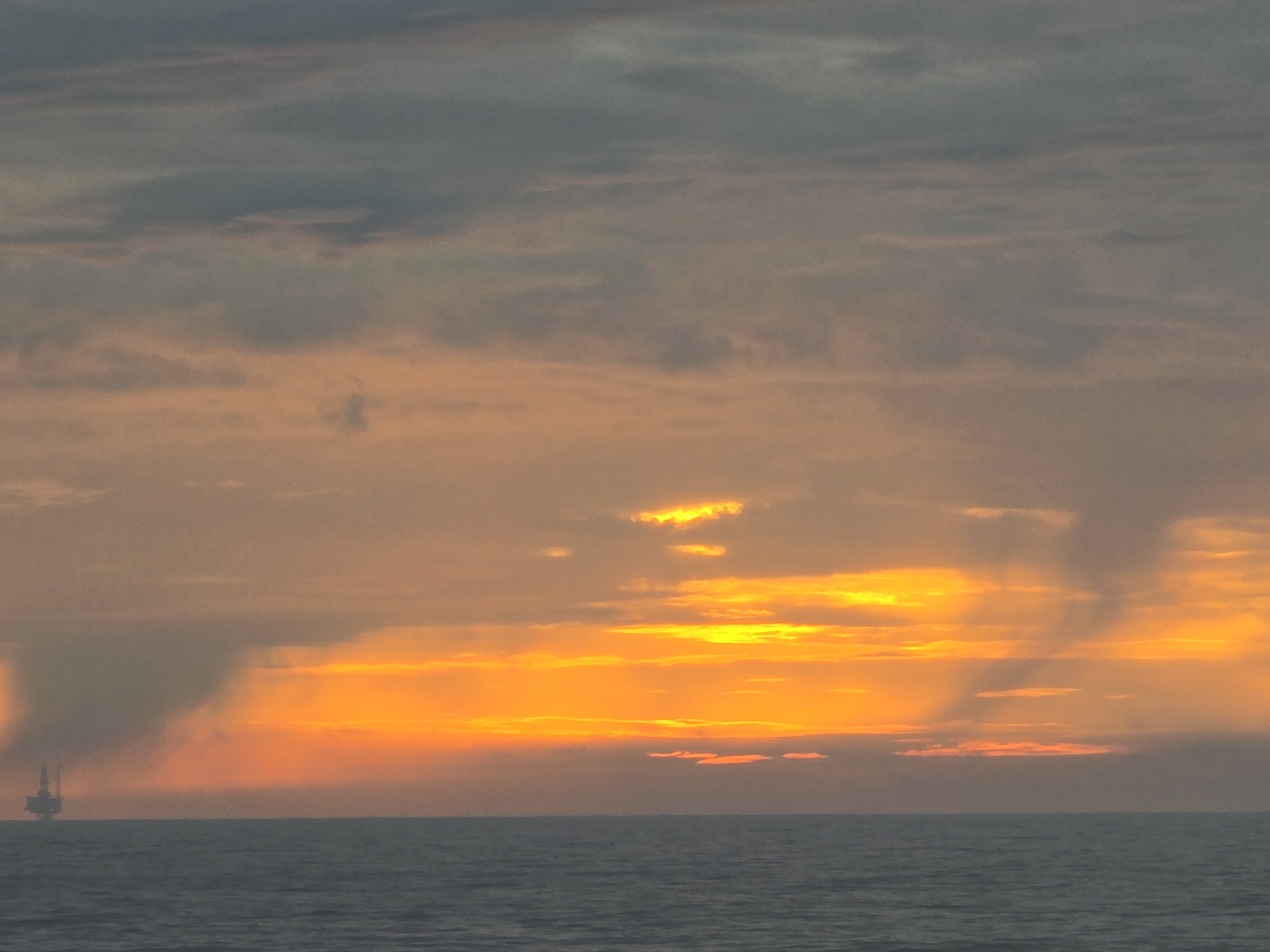 The image depicts an ocean scene at sunset, location unknown. The sun, a bright orange, is setting on the horizon, partially obscured by clouds. Darker clouds are present above the orange glow. A silhouette of what appears to be an offshore oil rig is visible on the left side of the horizon. The ocean water is gray-blue and relatively calm.
