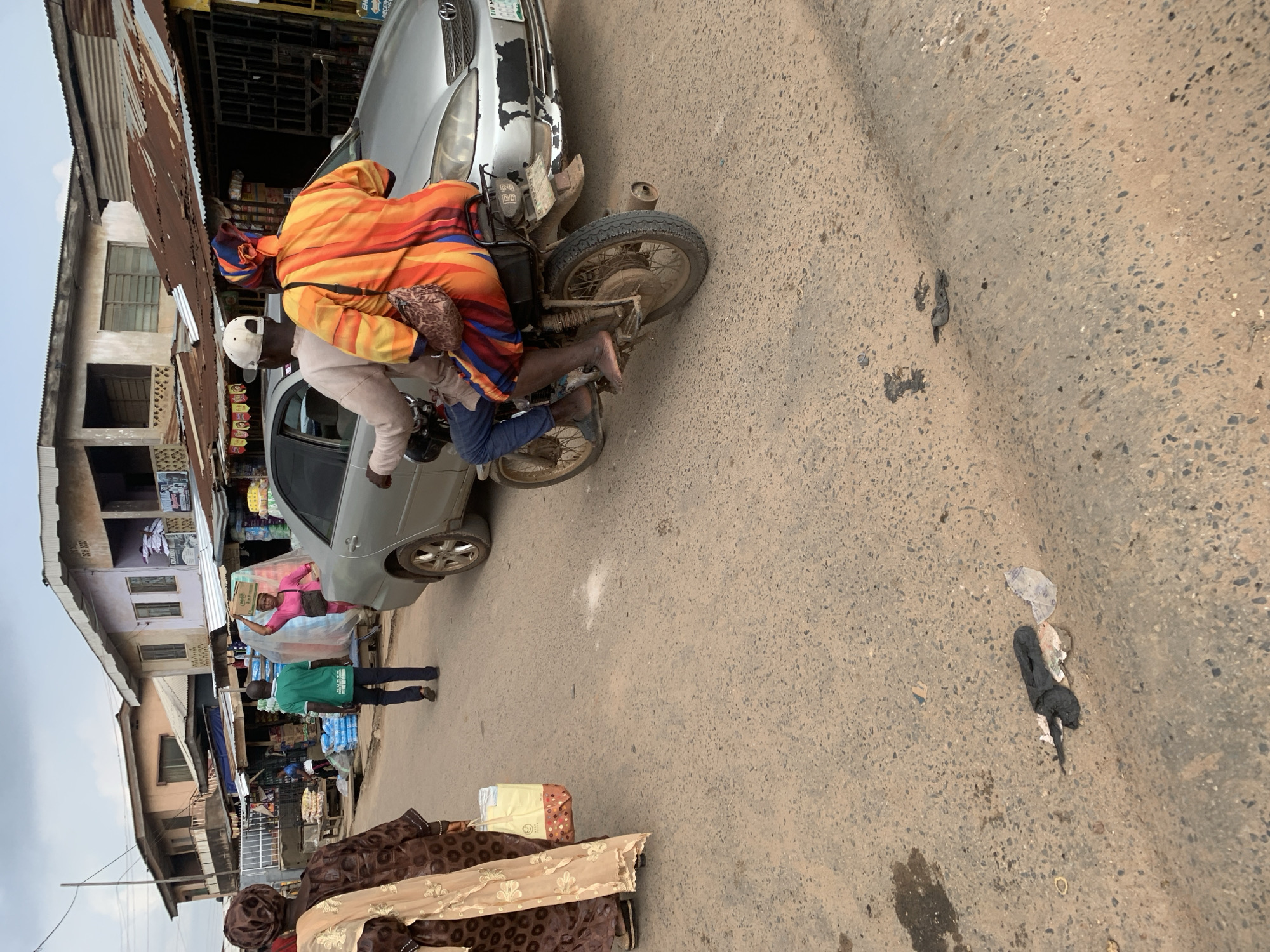 In a street scene in Shagamu, Nigeria, two people are on a motorcycle next to a silver car. The person in front, wearing an orange, yellow, and blue striped garment and a headwrap, is seated behind the driver. The driver, wearing a light-colored shirt and blue jeans, is wearing a white cap. The motorcycle is parked next to the car, which has a dented fender.
In the background, there are buildings that appear to be shops, with corrugated metal roofs and awnings. Some of the shops have goods displayed outside. A person in a pink shirt is seen standing next to the car, carrying a box. Another person in a green shirt is standing on the street. The ground is paved with asphalt and appears somewhat worn. There is some scattered debris on the street, including plastic bags and what appears to be animal waste.
The image appears to have been taken during the day, with overcast or cloudy conditions suggested by the lighting. The scene depicts a typical street in a busy commercial area, possibly a market, with people engaged in daily activities. The overall atmosphere seems busy and functional. There is no visible text that can be clearly read.