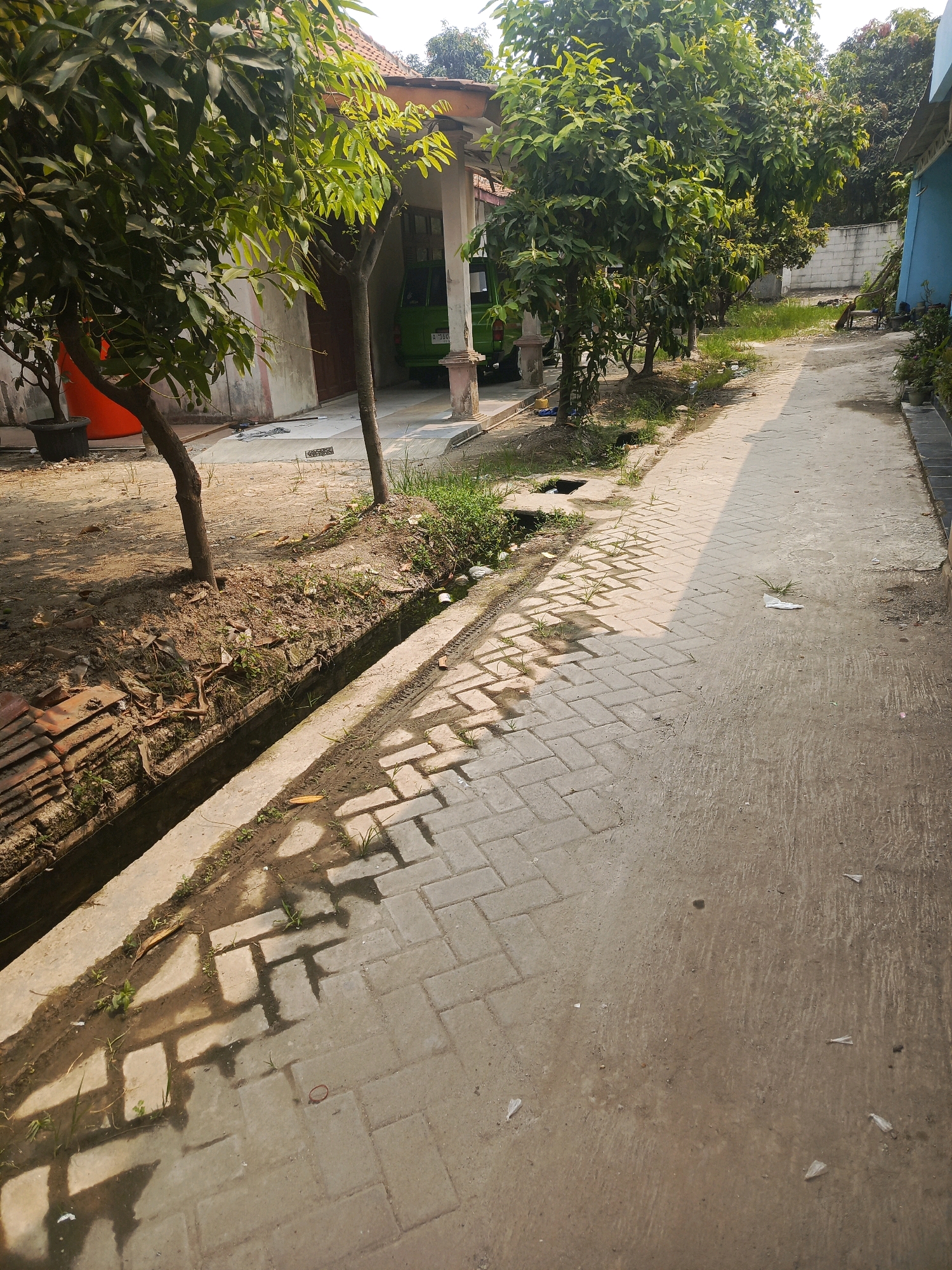 The image depicts an outdoor scene on a sunny day, likely in a residential area of Balaraja, Indonesia. A paved walkway, composed of rectangular bricks laid in a herringbone pattern, runs diagonally across the frame. To the left of the walkway is a shallow, open drainage ditch, partially filled with water and debris, bordered by dry soil and sparse vegetation. Several trees with lush green foliage are visible along the edge of the walkway and the property to the left, casting shadows on the pavement.
Adjacent to the trees on the left, part of a building with a light-colored wall and a tiled roof is visible. A large orange container and a black planter are also present in this area. Further down the walkway, on the right side, a lighter-colored, possibly concrete path runs parallel to the bricked section. Beyond this path, a light blue building is partially visible. In the background, a white brick wall and more trees are present. Small pieces of litter, including plastic wrappers, are scattered on the ground. The scene suggests a well-lit, possibly humid environment characteristic of Southeast Asia.