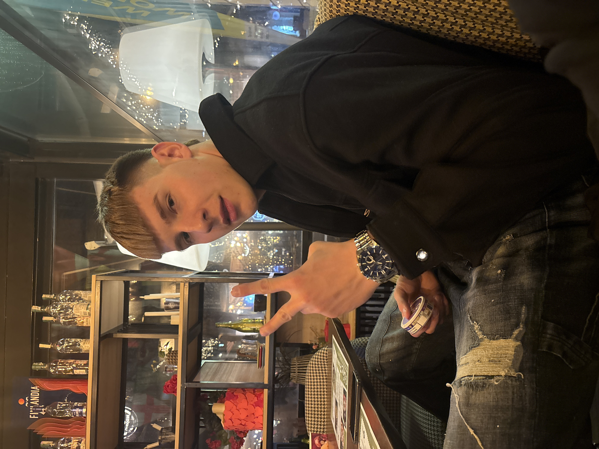 A young adult male is seated indoors, likely within a bar or restaurant environment in Sofia, Bulgaria, looking directly at the camera with a slight smile. He has short, light brown hair styled with a fringe and faded sides. He wears a black collared jacket and distressed blue jeans with visible tears on the left thigh. On his left wrist, he wears a silver-toned wristwatch with a blue dial. His right hand is raised, forming a two-finger gesture (index and middle fingers extended), commonly recognized as a "peace" or "victory" sign. His left hand holds a small, round tin with a purple label, consistent with nicotine pouch packaging.
The subject is seated on a chair or booth upholstered in a black and yellow/tan houndstooth or similar patterned fabric. A dark-colored table is positioned in front of him, displaying menus or papers and a smartphone.
The background features a multi-tiered display shelf unit to the left, prominently featuring multiple bottles of "FINLANDIA" vodka and a corresponding blue and red branded sign on the top shelf. The middle shelf contains decorative items including candles, a single red rose, and a small shot glass. The lower visible shelf holds an ornamental red item and a golden-colored bottle.
Behind the subject and the shelving unit, large windows reveal a blurred urban night scene with various colored lights, consistent with street lighting or decorative displays. Water droplets are visible on the exterior of the windowpane on the right side of the image. A white lampshade from inside the establishment is reflected in this window. The interior lighting is dim, contributing to an evening ambiance. The precise street address or specific name of the establishment in Sofia cannot be definitively identified from the provided visual information.