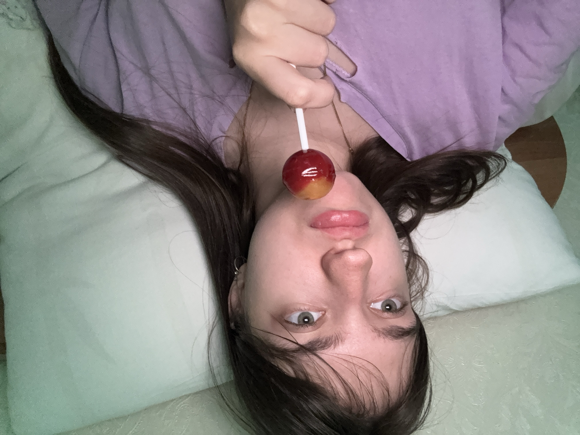 A young woman with light skin and dark hair is lying on a bed, holding a red and yellow lollipop in front of her nose. She is wearing a purple shirt and her hair is spread out on a white pillow. She has light eyes and visible eyebrows. The bed appears to be covered in a light green or white blanket. The photo is taken from an upside-down perspective. Location: Unknown, Unknown.