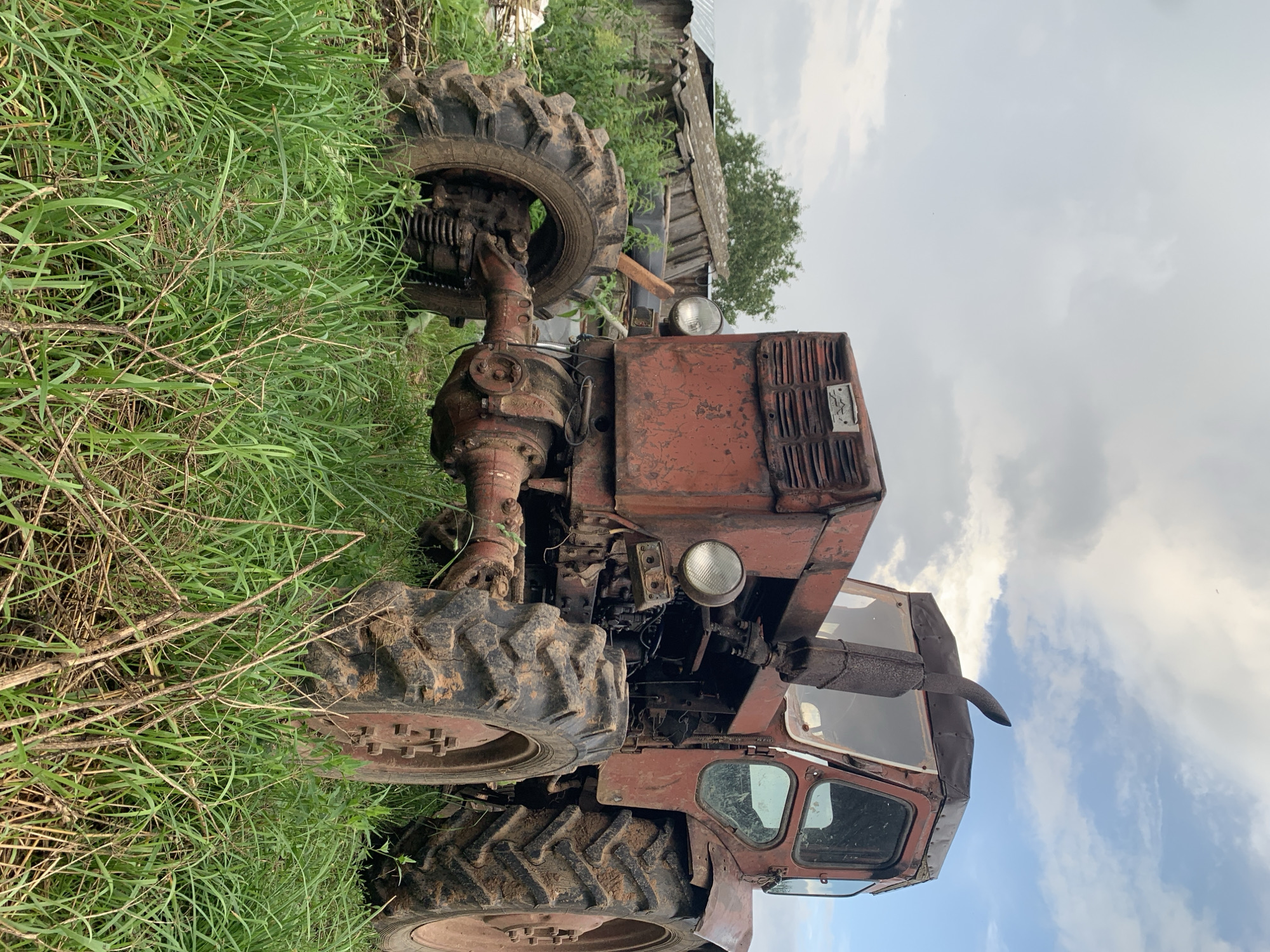 An old, weathered, red tractor is parked in a grassy field under a cloudy sky. The tractor is muddy, suggesting recent use in wet conditions. The front of the tractor is visible, showing its large, rugged tires, headlights, and a prominent exhaust pipe. In the background, a dilapidated wooden structure, possibly a shed or barn, leans precariously. Lush green grass surrounds the tractor, with some dry stalks and weeds interspersed. The location is identified as Panteleevka, Russia. There are no people or animals visible in the image. The scene appears to be rural and somewhat neglected.