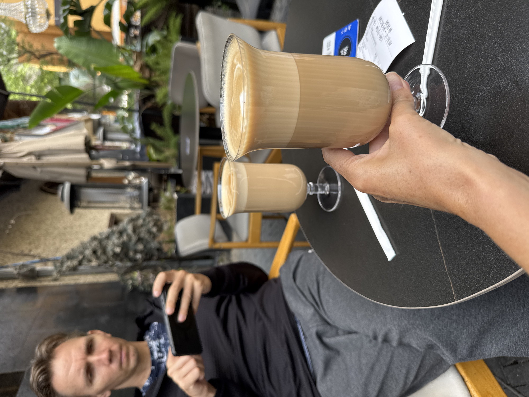 A person is holding a glass of coffee or a latte in their right hand over a round black table, while another person is seated and using a smartphone to take a picture. There are two drinks on the table, both appearing to be a similar type of coffee drink in clear glasses with a stem. A white straw and a bill are also on the table. The seated person is wearing a black shirt and gray pants. The location is Jinjiang District, China. In the background, there is greenery, chairs, and an outdoor dining area.