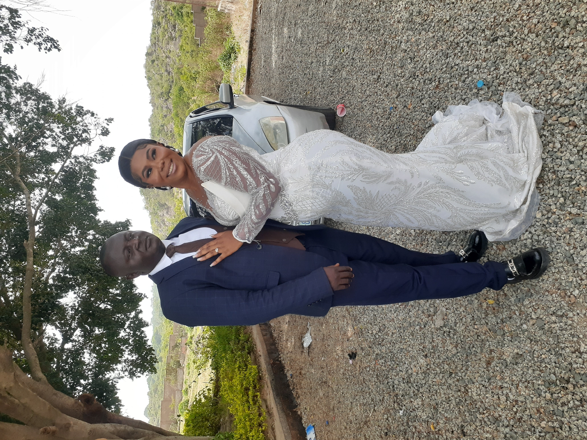 A full outdoor shot depicts a bride and groom posing in front of a silver car on a gravel surface, set against a background of trees, shrubs, and a distant mountain.
The bride, positioned above the groom, is smiling at the camera. She wears a long-sleeved, fitted white gown embellished with glittery designs. The groom, lying down, is also smiling. He wears a navy blue suit with a brown tie and shiny black shoes.
The location appears to be a rural area with natural vegetation and a mountain in the distance. A tree and shrubbery are visible in the foreground. A red cup is on the gravel near the car.
