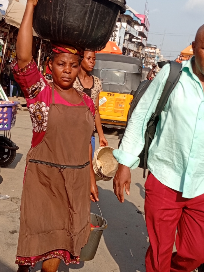 A woman carries a large black tub on her head in a bustling outdoor market. She wears a brown apron over a patterned red dress and holds a bucket. A young woman stands behind her. A yellow vehicle with "TVS" branding is visible. A man in a light green shirt and red pants walks beside her, carrying a backpack. The scene is lively and shows daily life.