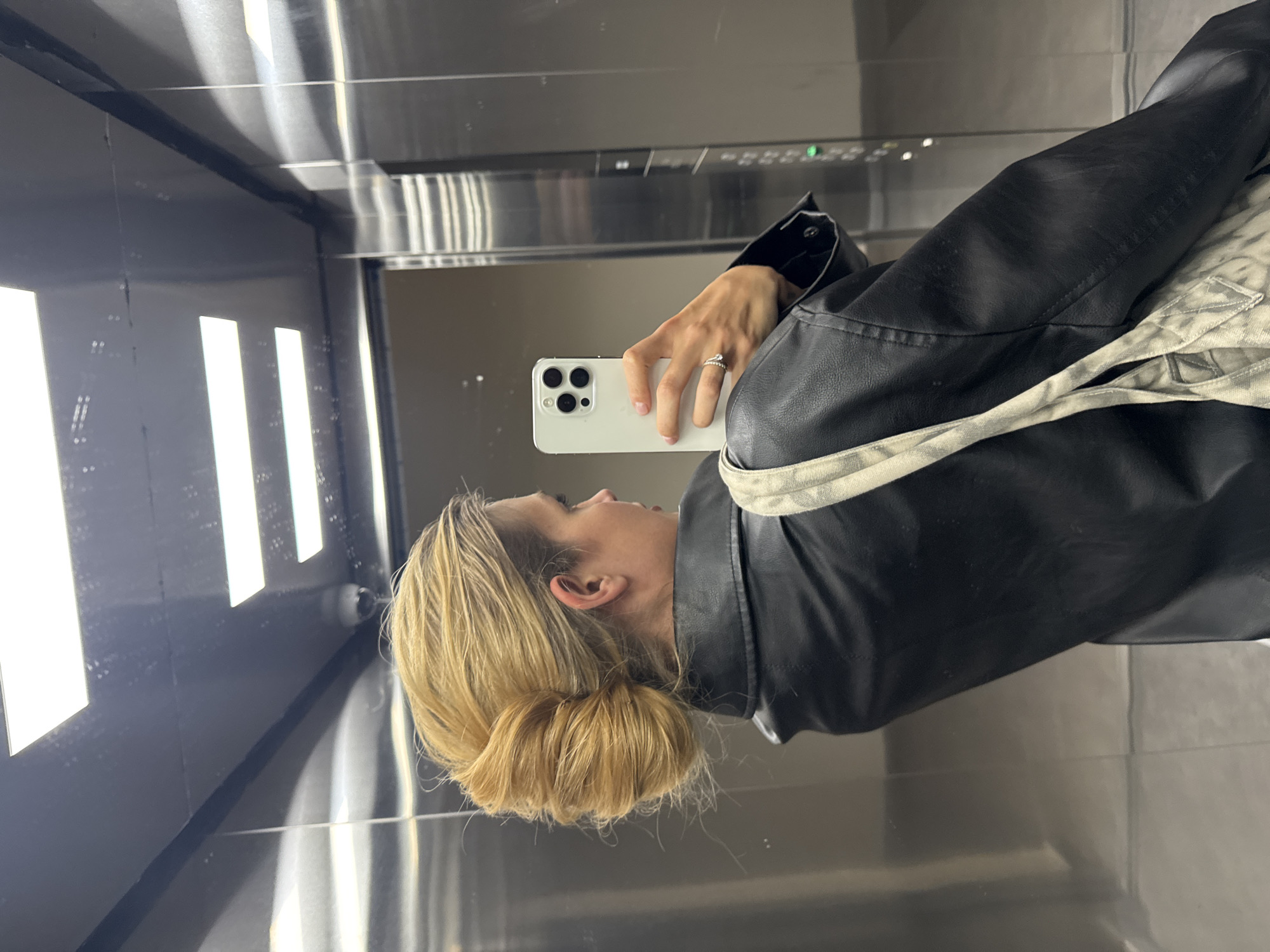 A woman is taking a selfie in an elevator. She is wearing a black leather jacket and has blonde hair pulled back in a bun. She is holding a white smartphone in her right hand, which is adorned with a ring. The elevator has stainless steel walls with vertical light panels. There are elevator buttons visible in the reflection of the walls. The location is Unknown, Unknown.