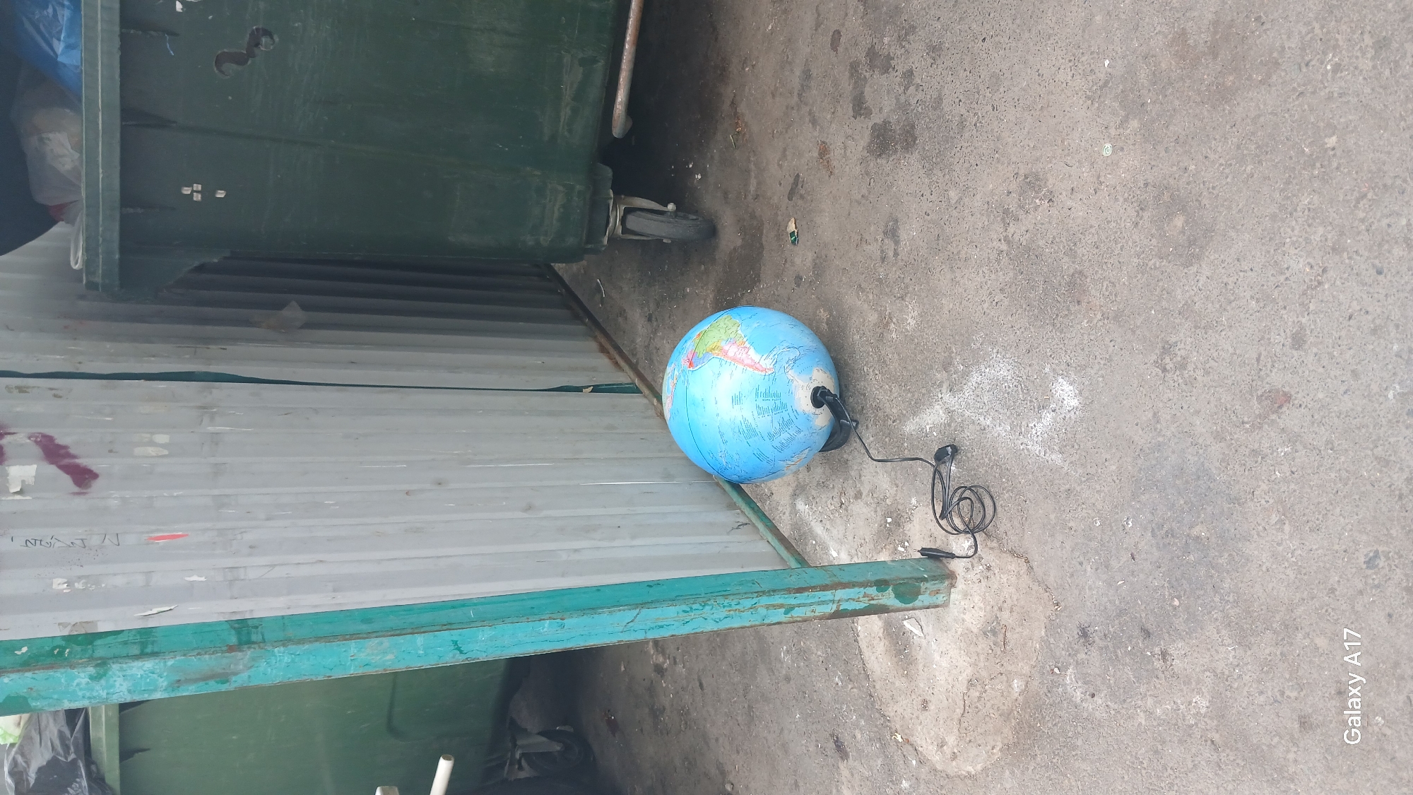 The scene is an outdoor urban environment, possibly an alleyway or service area in Yekaterinburg, Russia, judging by the Cyrillic lettering visible on the gray corrugated metal wall. The scene contains:
- A partially visible green dumpster on the left and top of the frame.
- A gray corrugated metal wall.
- A blue globe with geographic markings with a black cord plugged into it. The cord is coiled on the gray concrete ground.
- Gray concrete ground.
- The words "Galaxy A17" in the bottom right corner.