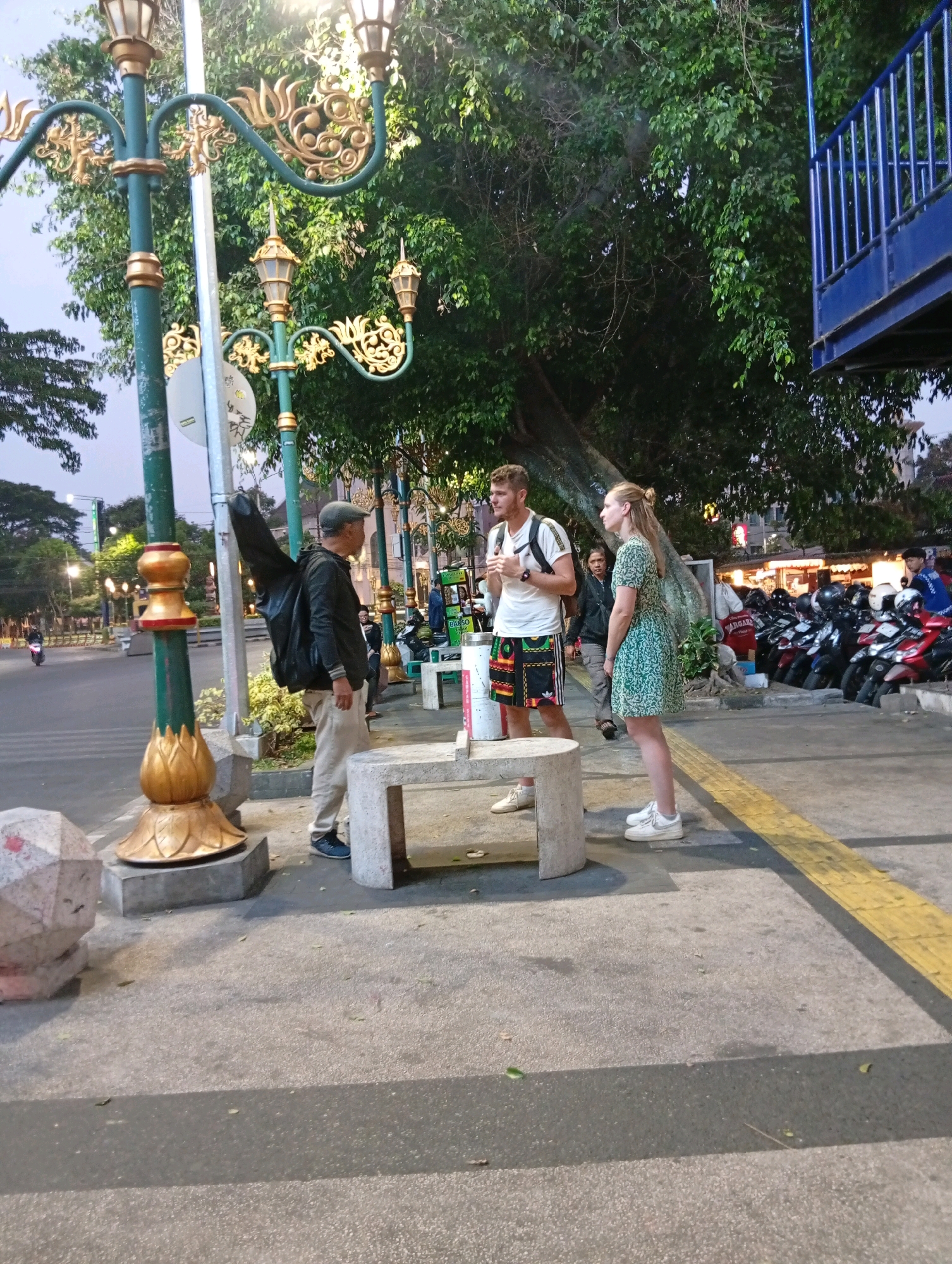 The image depicts an outdoor scene on a city street, possibly in Malang, Indonesia, with a focus on pedestrians and street elements. In the foreground, a man wearing a cap, dark jacket, and light-colored pants stands with a black guitar case slung over his back. He is facing away from the camera and to the right. Next to him, a man with blonde hair and a white t-shirt and patterned shorts is talking to a woman with blonde hair wearing a green patterned dress. They are standing near a concrete bench. Behind them, a line of parked motorcycles is visible, suggesting a busy urban environment. The background features lush green trees and ornate street lamps, indicating a park-like or well-decorated public space. The overall impression is of a casual encounter or waiting area in a lively part of the city.