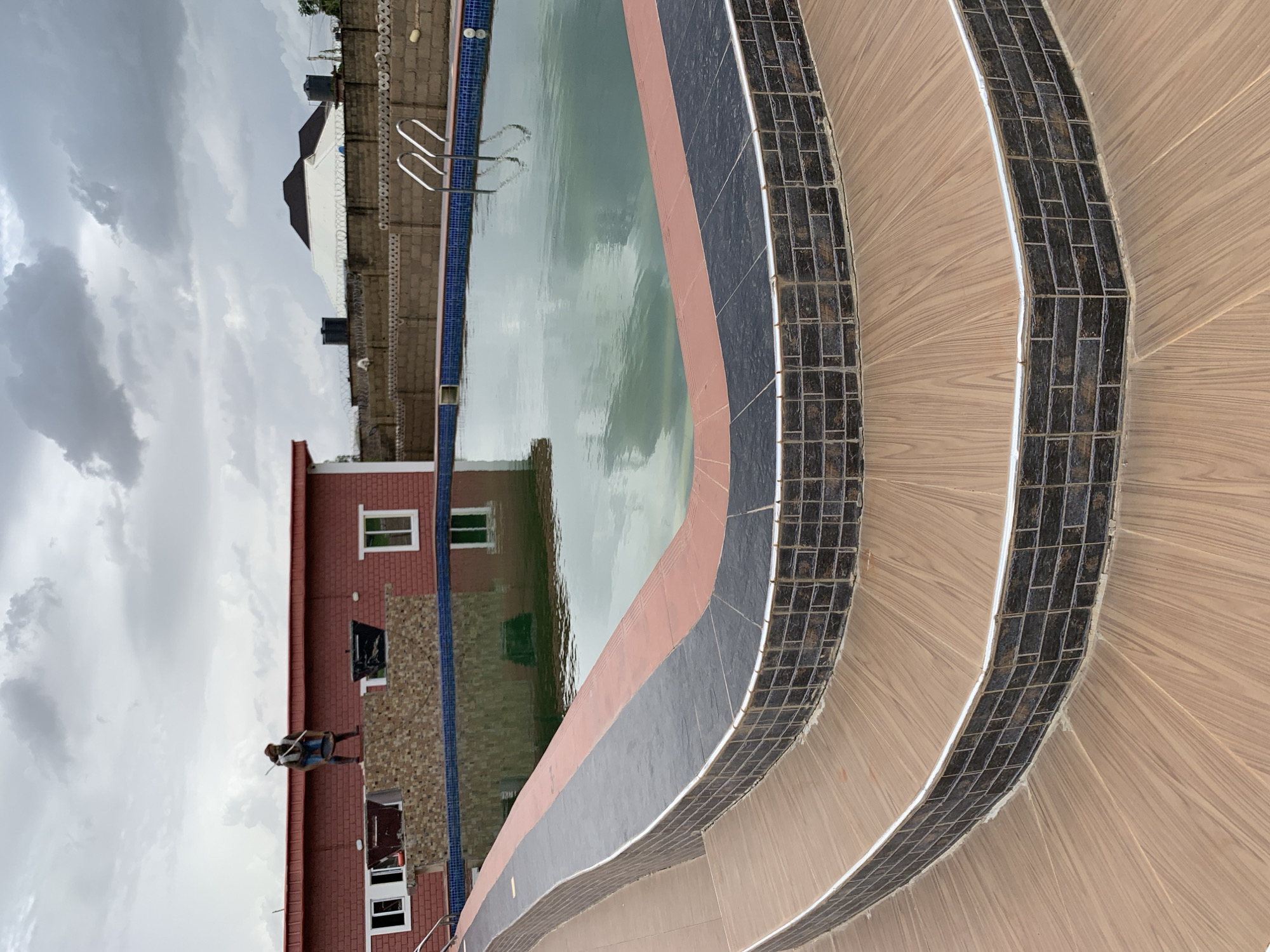 The image shows a swimming pool area on a cloudy day in Jos, Nigeria. In the foreground, a curved staircase with wooden steps and dark tiled accents leads down towards the pool. The pool itself is filled with green water and has a tiled edge. One person is visible near the edge of the pool, standing on the tiled area and appearing to hold a long pole or stick.
In the background, a red brick building with white-framed windows is situated next to the pool. Beyond the building, a brown brick wall topped with barbed wire suggests a perimeter fence. Several black water tanks are visible on top of the building and on the other side of the fence. The sky is overcast with heavy, gray clouds, indicating a potentially rainy or stormy weather condition. The overall scene suggests a residential or hospitality setting, possibly a private home or a small hotel. There is no visible text in the image.