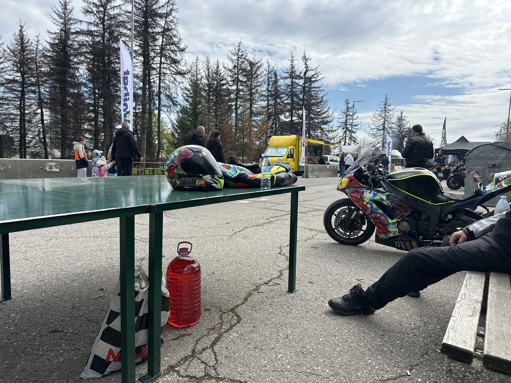 The image presents a scene at an outdoor event, possibly a motorcycle race or gathering, set against a backdrop of pine trees and a partly cloudy sky. Several individuals are present: some are standing and conversing in the background, while one person is seated on a bench in the foreground.
Key elements include a green metal table on which a motorcycle helmet and racing suit are placed. Beside the table, there's a bag and a large transparent bottle filled with a red liquid. A motorcycle, adorned with colorful graphics, is parked nearby.
The location is identified as Sofia, Bulgaria. The general atmosphere suggests a casual, possibly competitive, event.