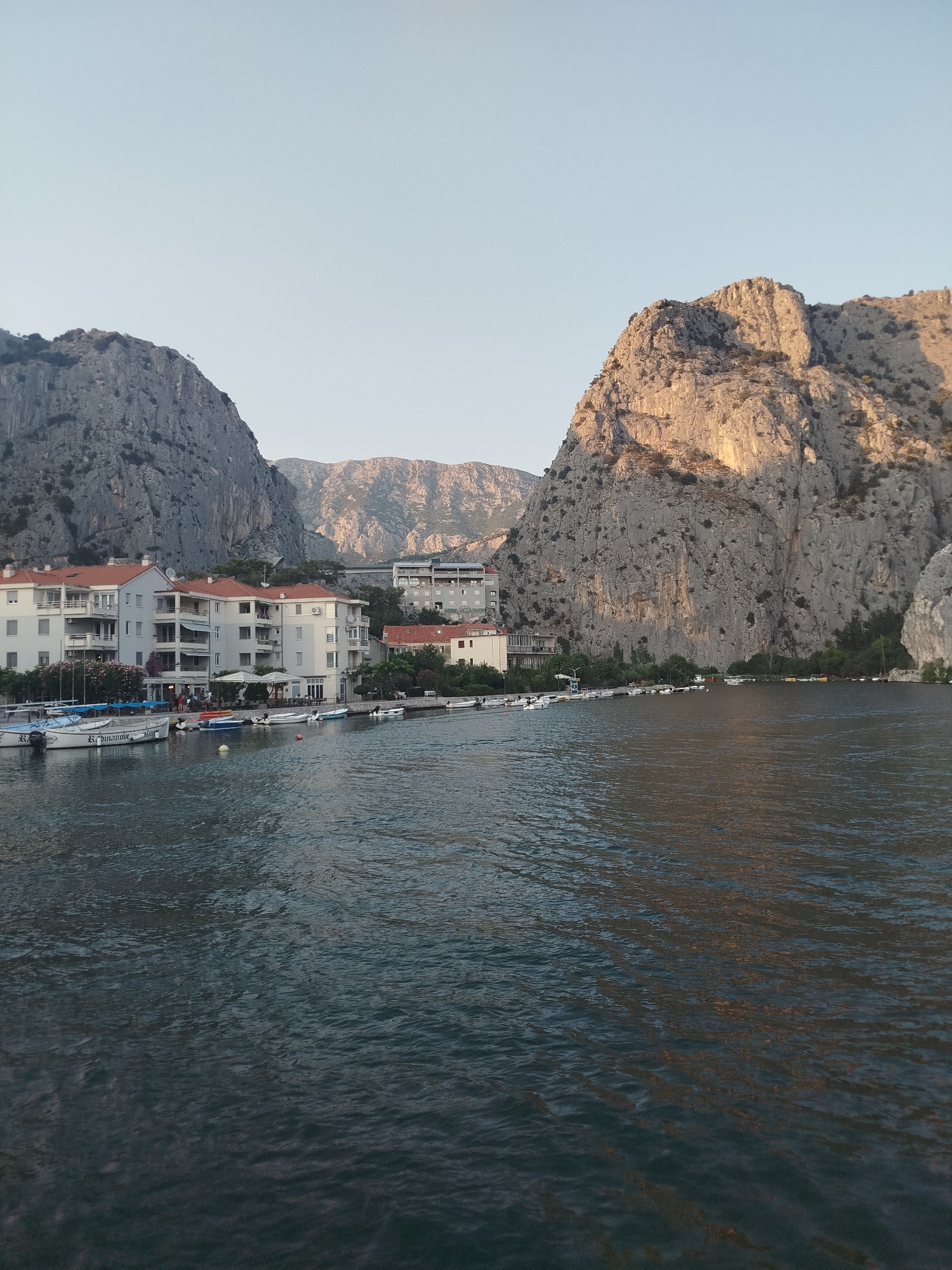 The image captures a scenic view of the Cetina River in Omiš, Croatia, with towering limestone cliffs framing the town. In the foreground, the dark water of the river flows, reflecting the sky and the surrounding landscape. Along the riverbank, a row of white buildings with terracotta roofs lines the waterfront. Several boats, including smaller fishing vessels and white motorboats, are moored along the shore. People can be seen gathered outside buildings, some sitting at outdoor tables, suggesting a leisurely atmosphere. The cliffs on either side of the river are imposing, with a scattering of vegetation clinging to their rocky surfaces. The right cliff face is illuminated by the warm glow of the setting sun, highlighting its rugged texture. In the background, more mountains are visible, stretching into the distance under a clear, pale blue sky. A bridge is subtly visible in the far background, connecting parts of the town.