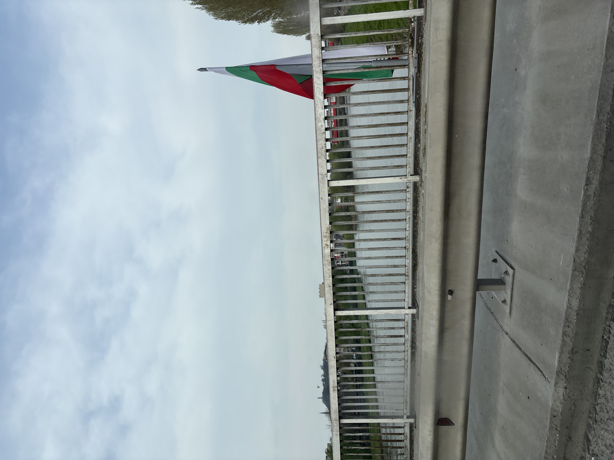 The image shows the Bulgarian flag attached to a bridge. The flag is composed of white, green, and red stripes. The bridge has a metal railing and a concrete barrier. The sky is cloudy and grey. Reflected in the water below is greenery and buildings, suggesting an urban location. The city is Unknown.
