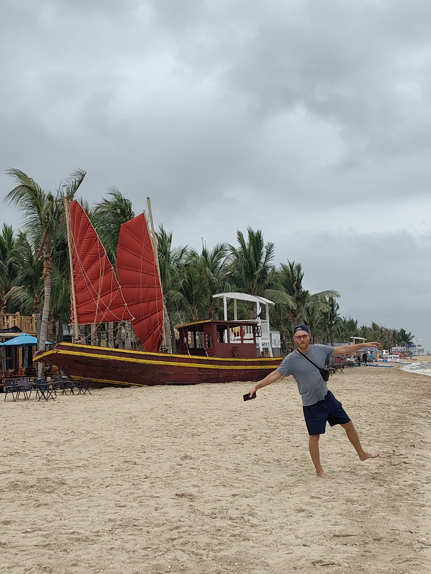 A man with a blue baseball cap, glasses, a gray shirt, and navy shorts stands barefoot on a sandy beach in Bai Chay Ward, Vietnam. He is balancing on one leg with his arms outstretched. He is holding a phone in his left hand. Behind him, there is a large, maroon-colored boat with red sails. The boat is surrounded by palm trees. The sky is overcast. There are chairs and tables visible to the left of the boat, suggesting a beachside establishment. Other people and boats are visible further down the beach.
