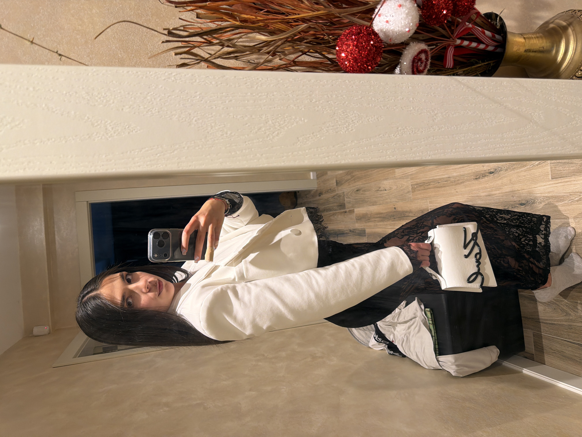 A young woman is taking a selfie with a dark grey smartphone. She is wearing a white blazer and a black lace dress. She has long, dark hair. To her right is a black box with white clothes piled on top, and a white purse with the word "Love" written on it. The floor is light brown wood. Behind her is a doorway leading into a darker room. On a shelf above is a decorative arrangement of branches and red and white ornaments in a gold vase. It is not possible to tell the location.