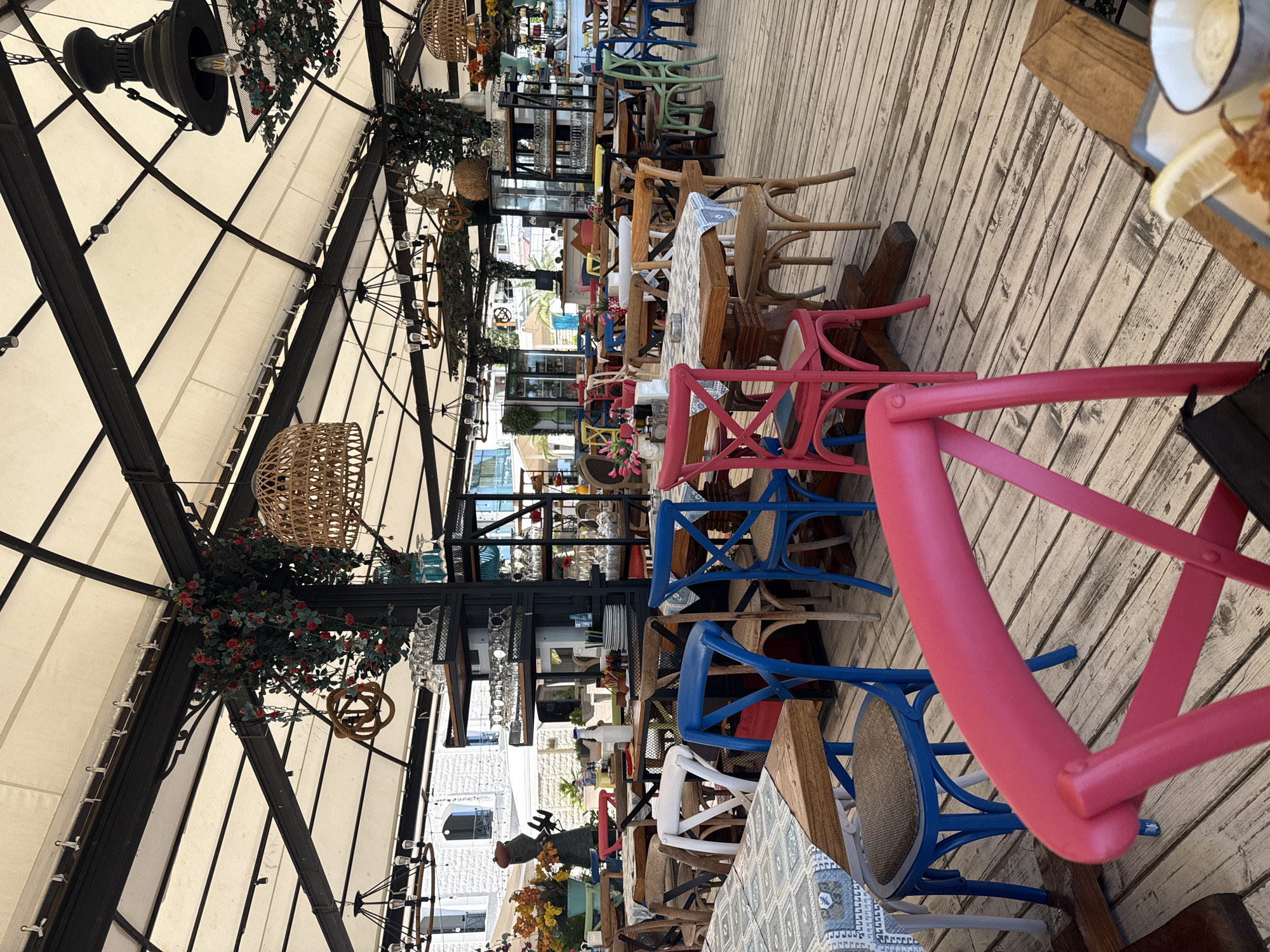 This is an outdoor dining area with wooden floors and a white canvas roof supported by black metal beams. The area is filled with tables and chairs of various colors, including blue, pink, and natural wood. Some tables are set with placemats and tableware. In the background, there are shelves with glassware and plates, and a glimpse of buildings with white stone walls. Decorative elements include hanging light fixtures, woven lampshades, and artificial red flowers draped from the beams. A person is partially visible in the distance, and a plate of food with a lemon wedge and a dipping sauce is in the foreground.